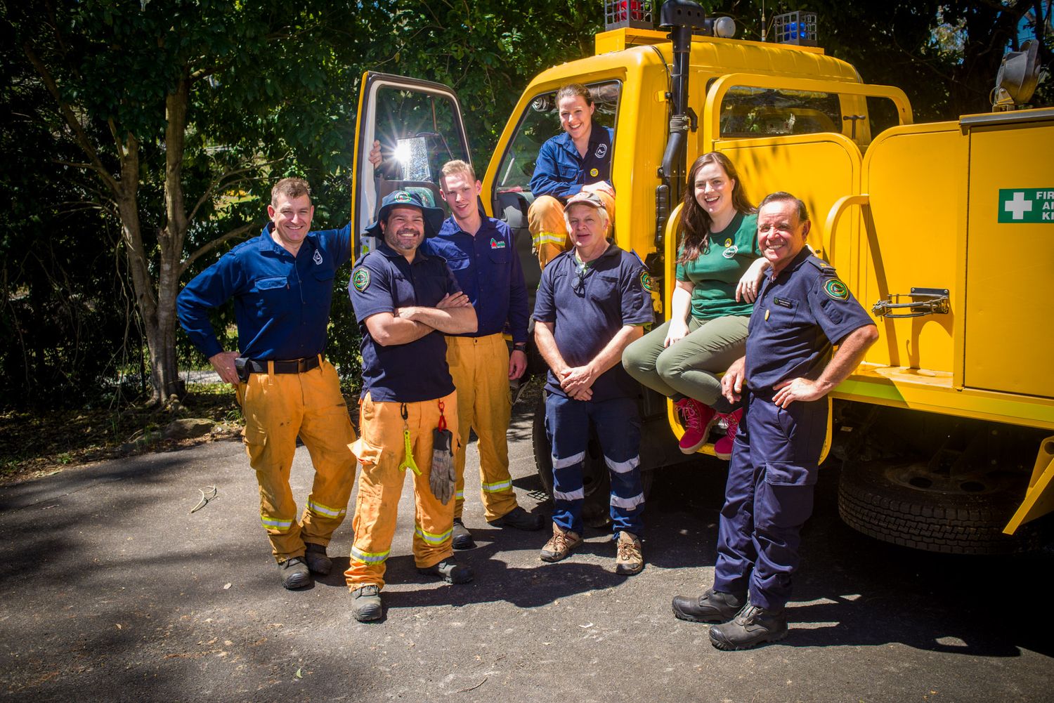 Group of seven emergency service workers posing beside a yellow fire truck outdoors in summer.
