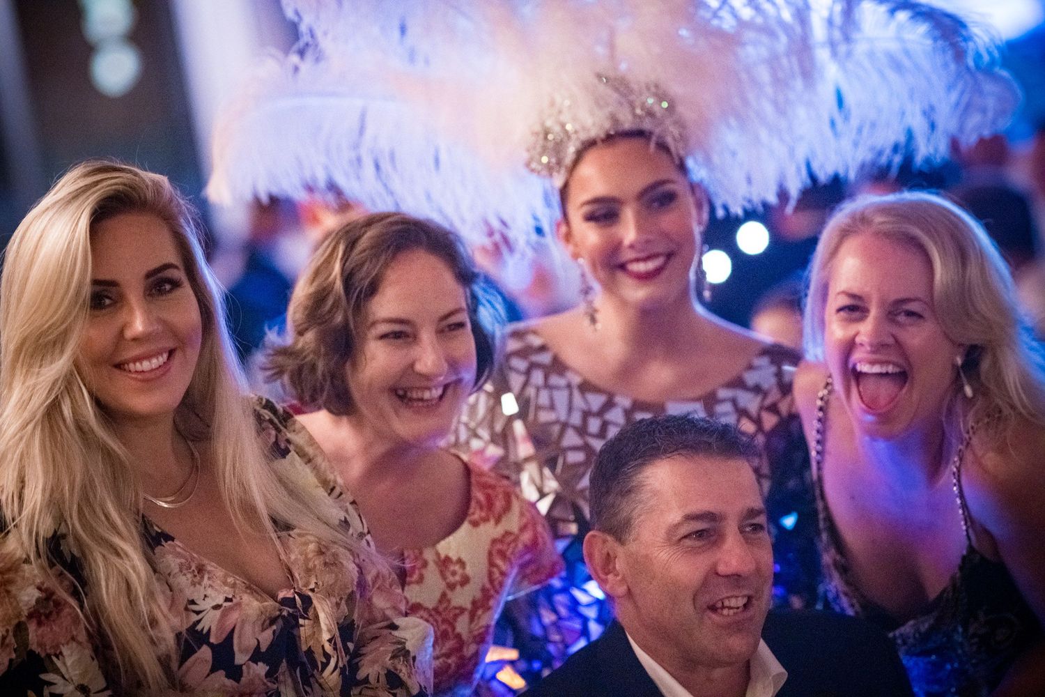Group of smiling people posing together at a glamorous gala event with feathered decorations.