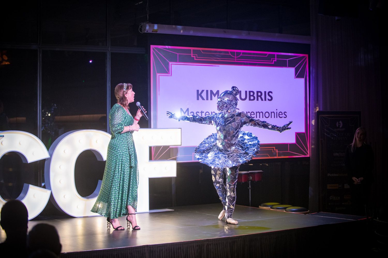 Female MC in green dress presenting award on stage with silver costumed performer at gala event.