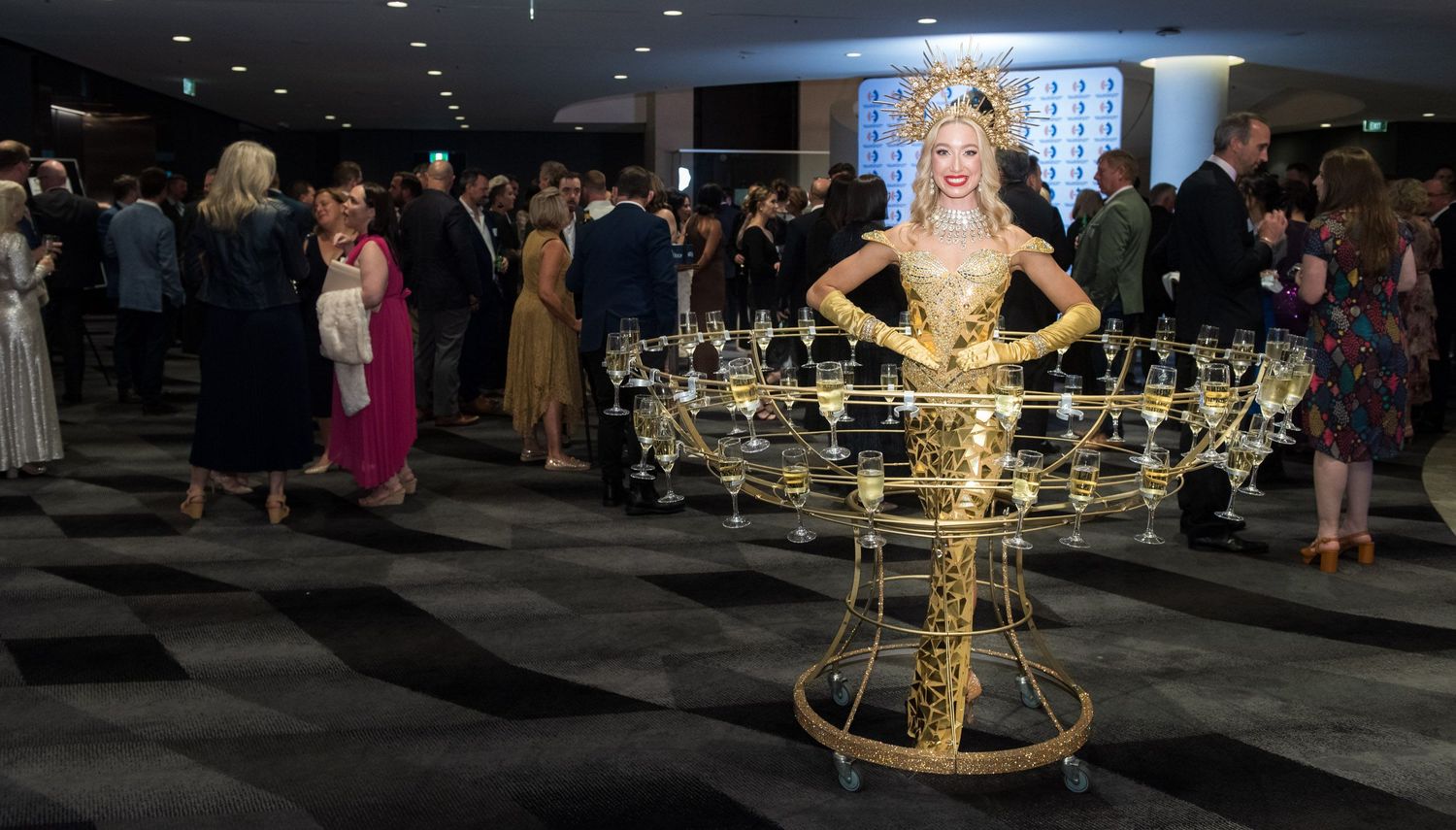 Elegant woman in gold costume carries champagne glass tray at upscale gala event.