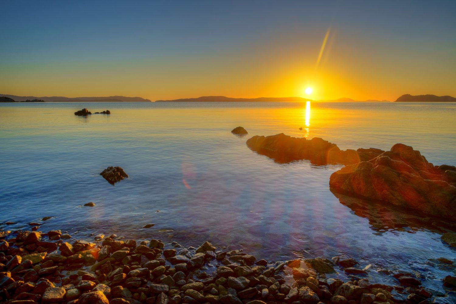 Golden sunset over calm rocky ocean coastline with warm reflections on still water.