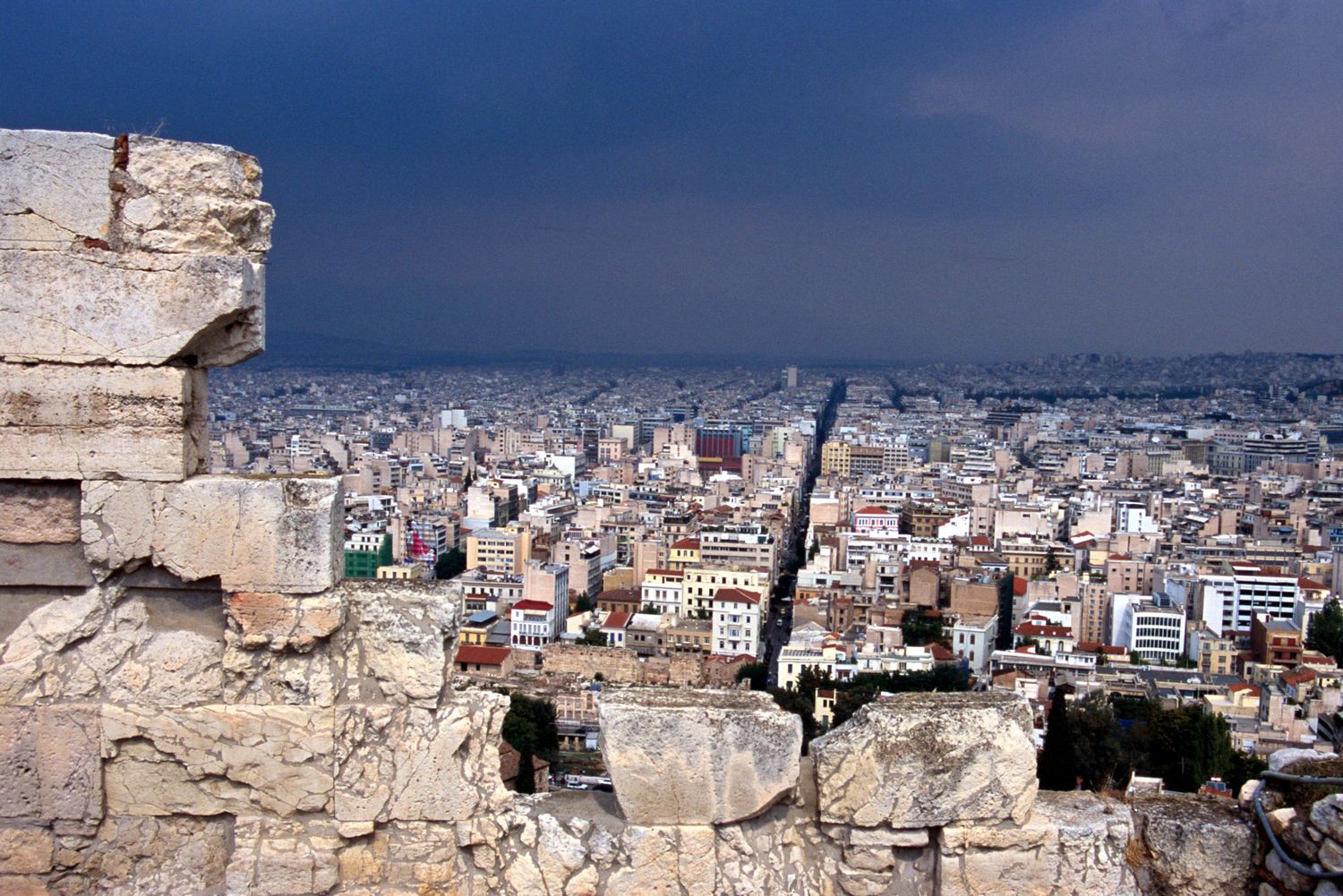 Panoramic view of Athens cityscape seen through ancient stone ruins under stormy skies.