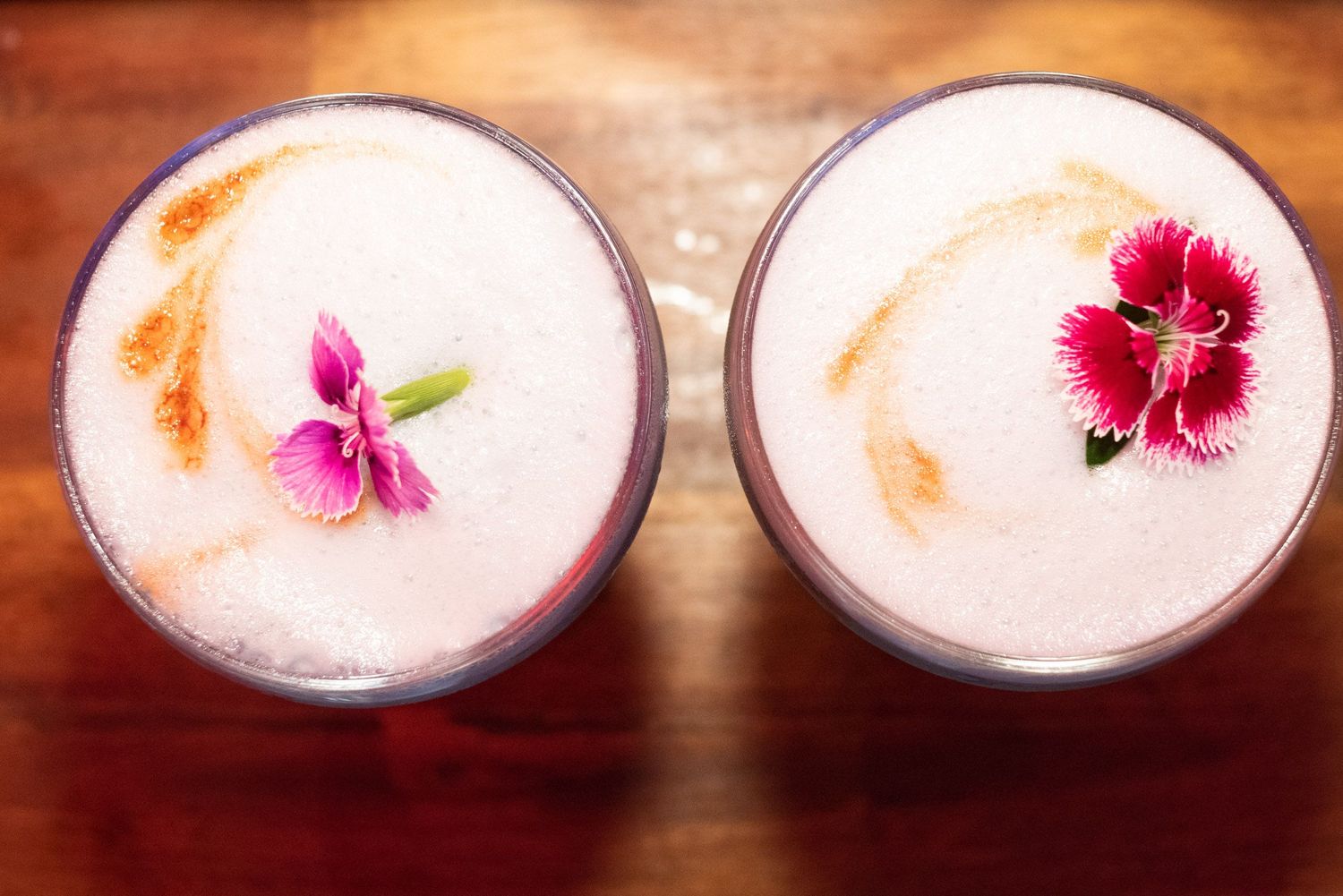 Two frothy cocktails garnished with pink carnation flowers on a warm wooden surface.