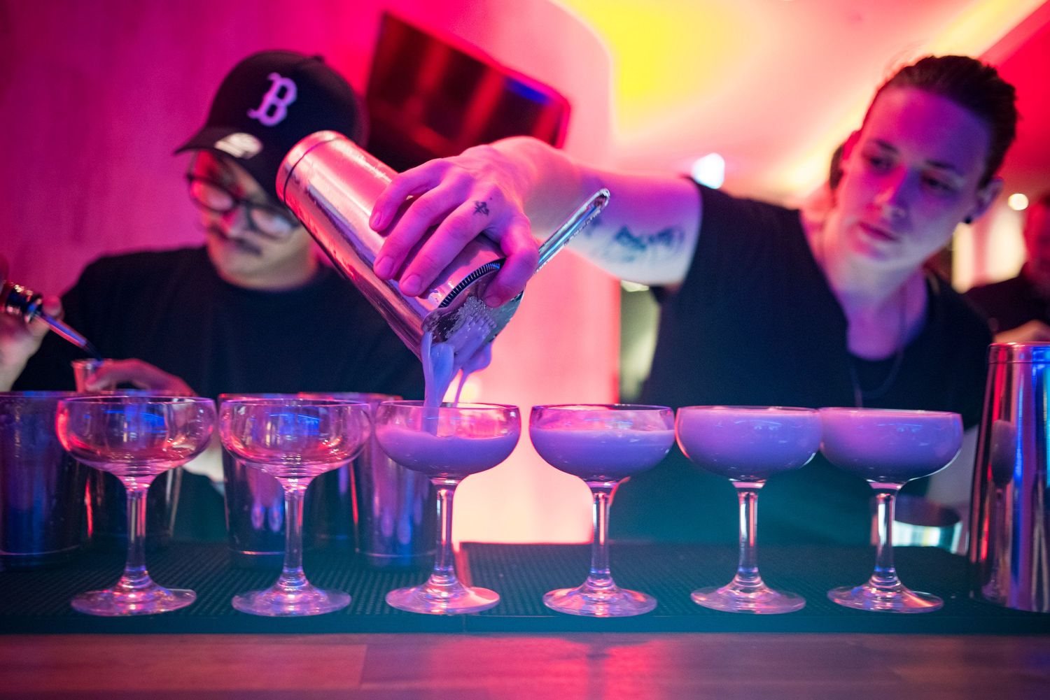 Bartender pouring cocktails into row of elegant coupe glasses under pink and blue neon lights.