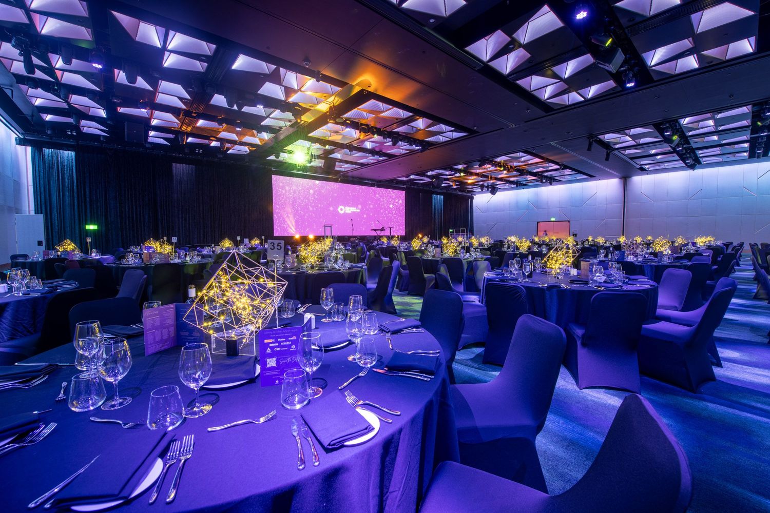 Elegant ballroom gala setup with purple lighting, round tables, geometric centerpieces, and large screen backdrop.