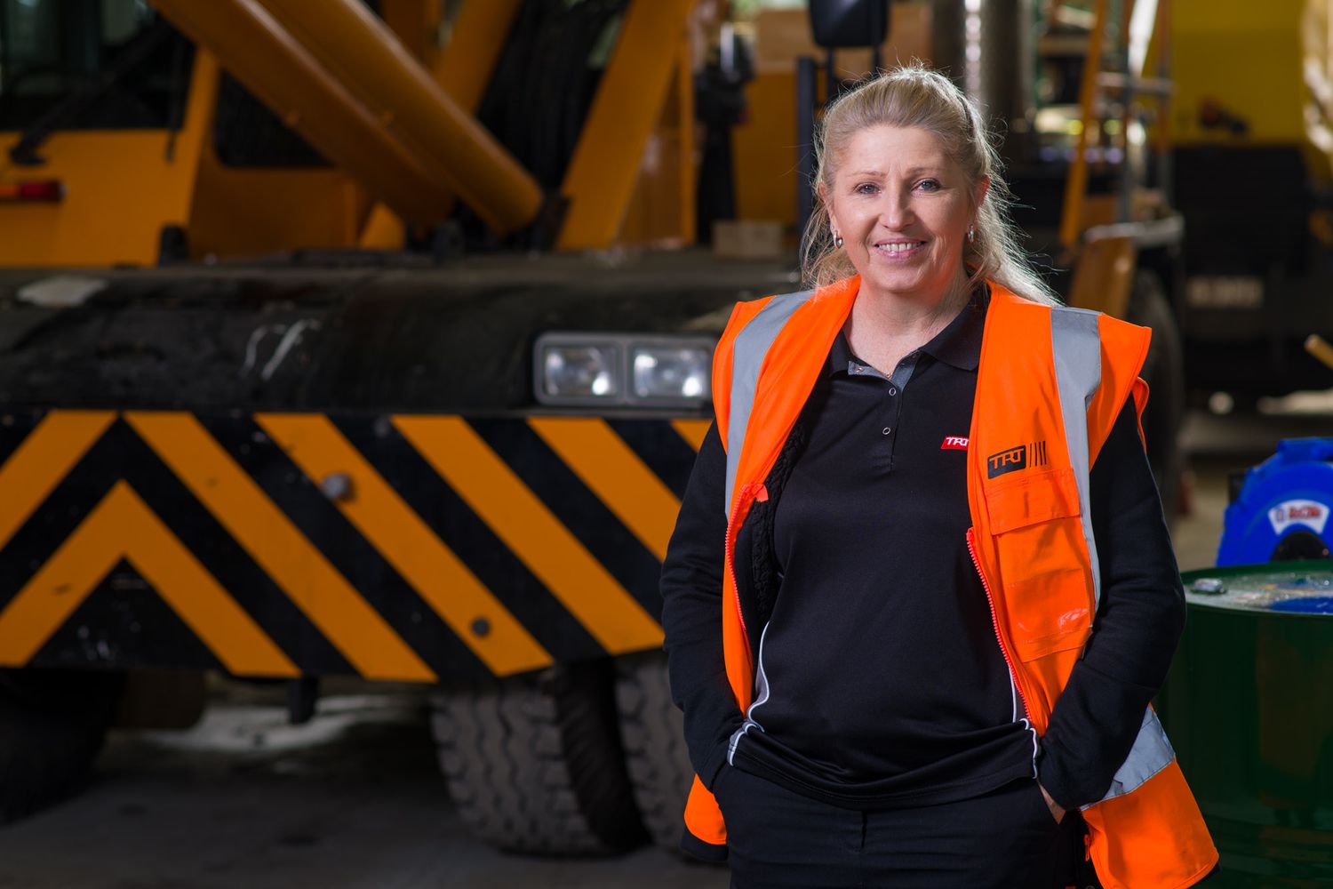 Woman in orange hi-vis vest standing confidently in front of heavy construction equipment.