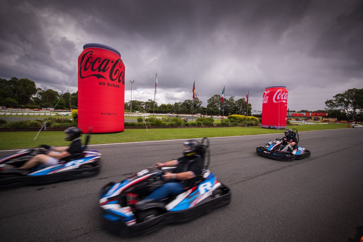 Three go-karts race past giant inflatable Coca-Cola cans on an outdoor track under stormy skies.