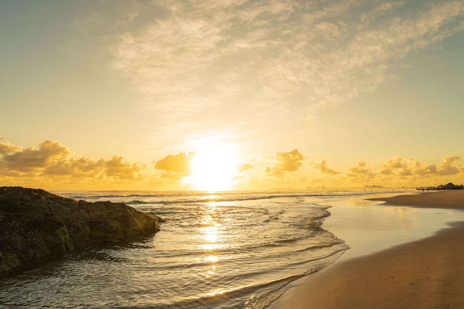 Golden sunrise over a serene beach with gentle waves, rocks, and glowing reflections on wet sand.