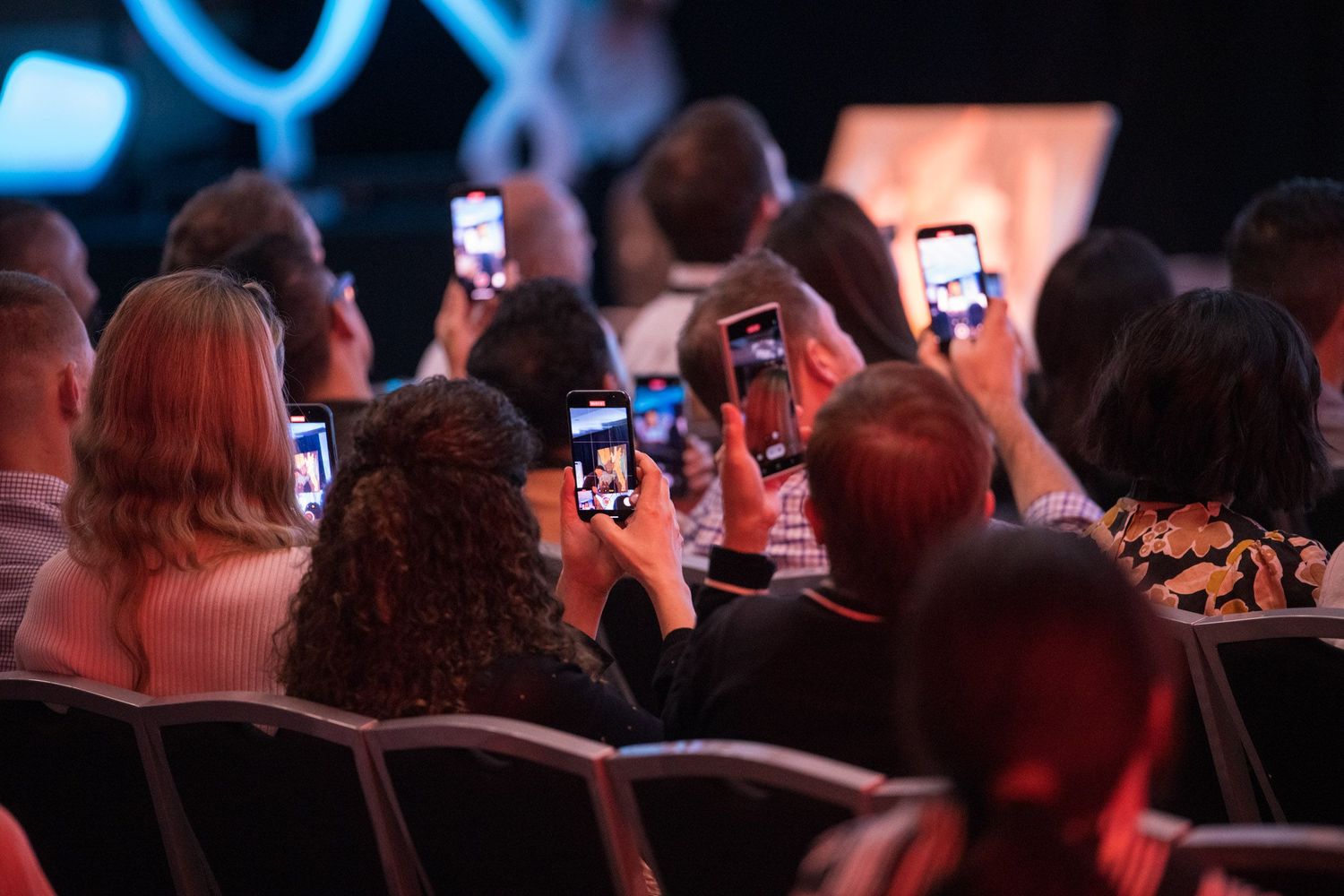 Conference audience members photograph a stage presentation using smartphones in a darkened venue.