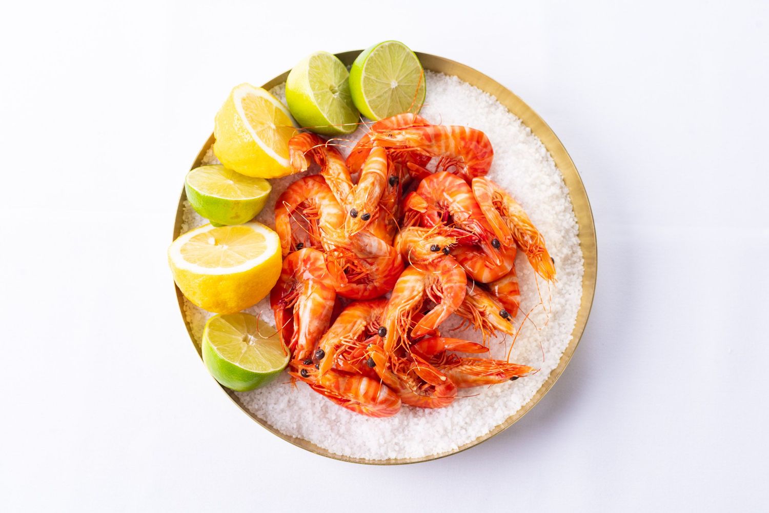 Cooked prawns served on a salt bed with lemon and lime wedges on a round plate.