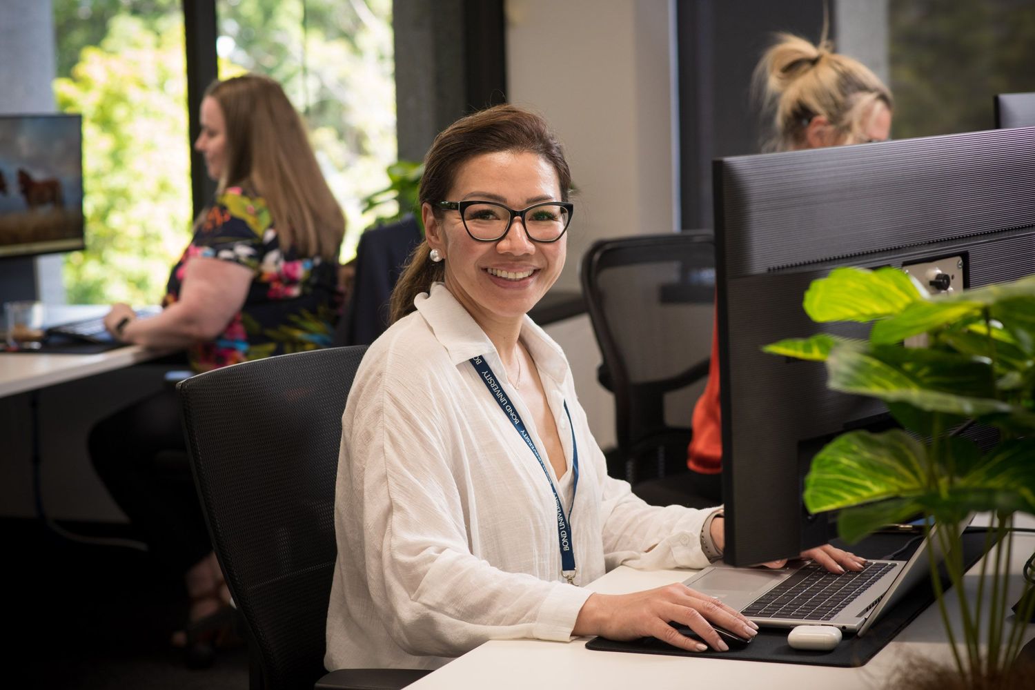 Smiling woman with glasses works at a computer desk in a bright open-plan office with green plants nearby.
