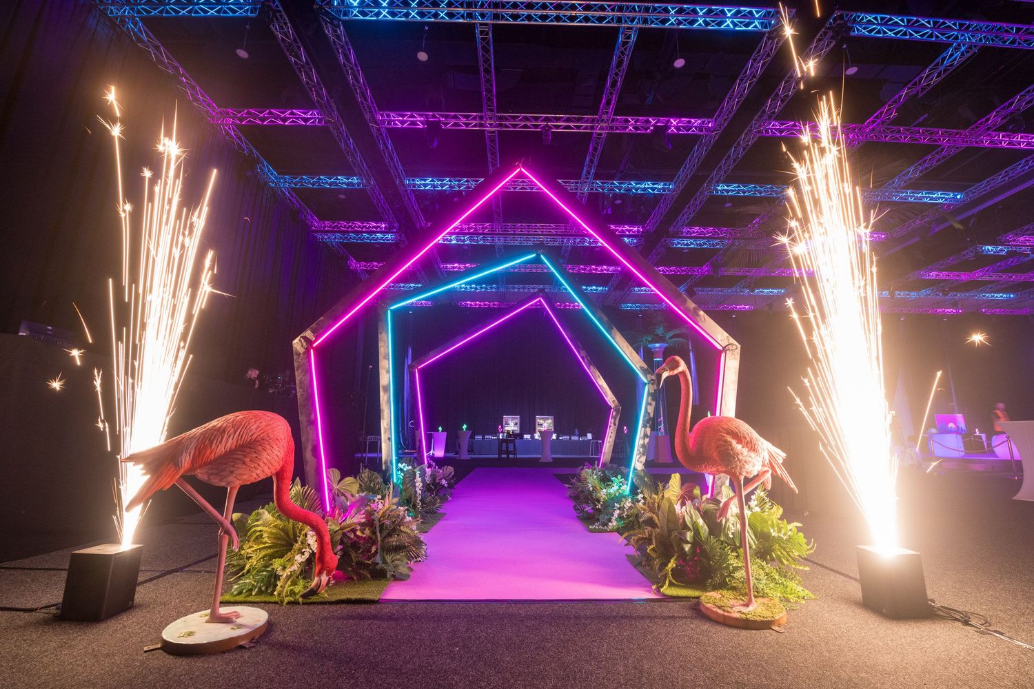 Spectacular event entrance with neon geometric arches, flamingo sculptures, and pyrotechnic sparklers on purple carpet.