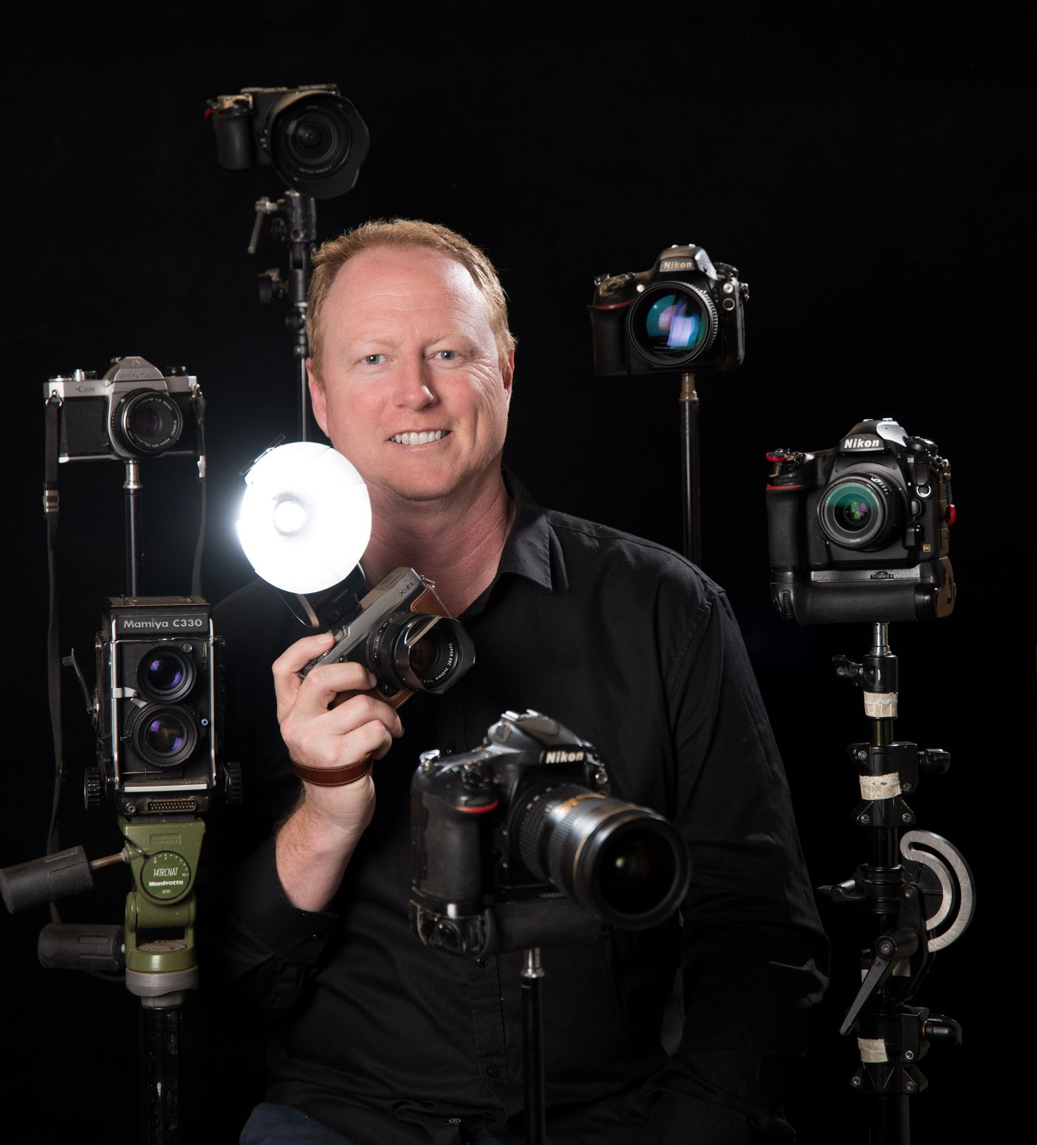 Professional photographer surrounded by multiple cameras and equipment on black background.