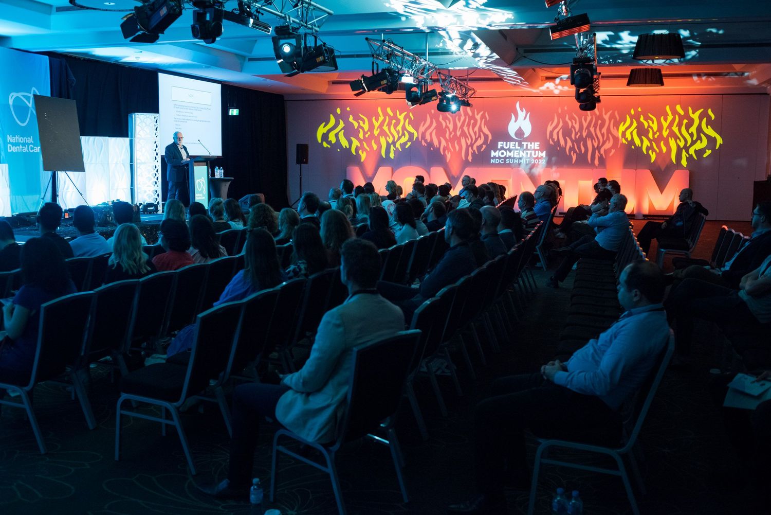 Audience seated in darkened conference hall watching a dramatic fire-themed stage presentation.