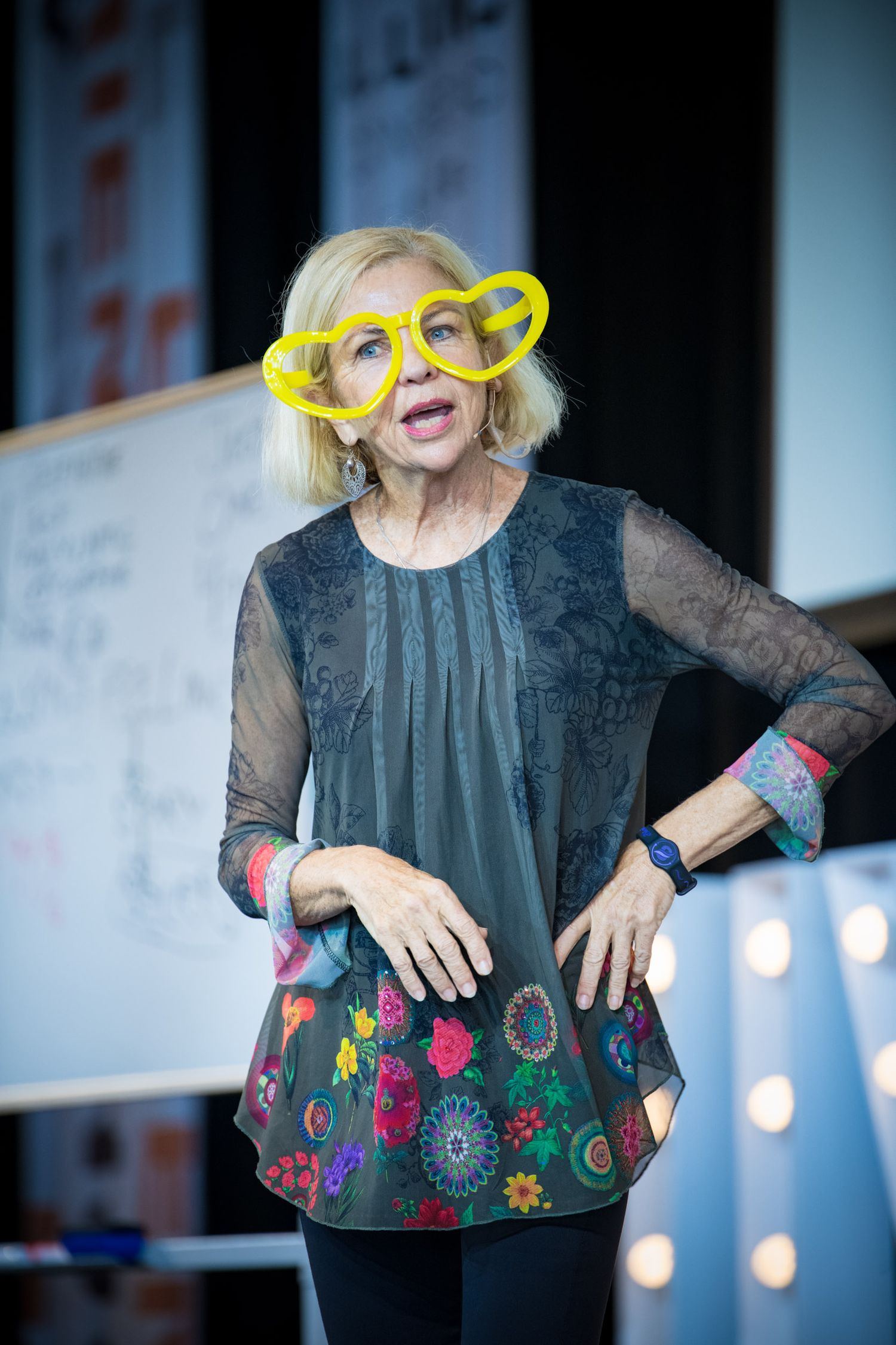 Woman wearing oversized yellow heart-shaped glasses and floral tunic speaks on stage at a conference event.