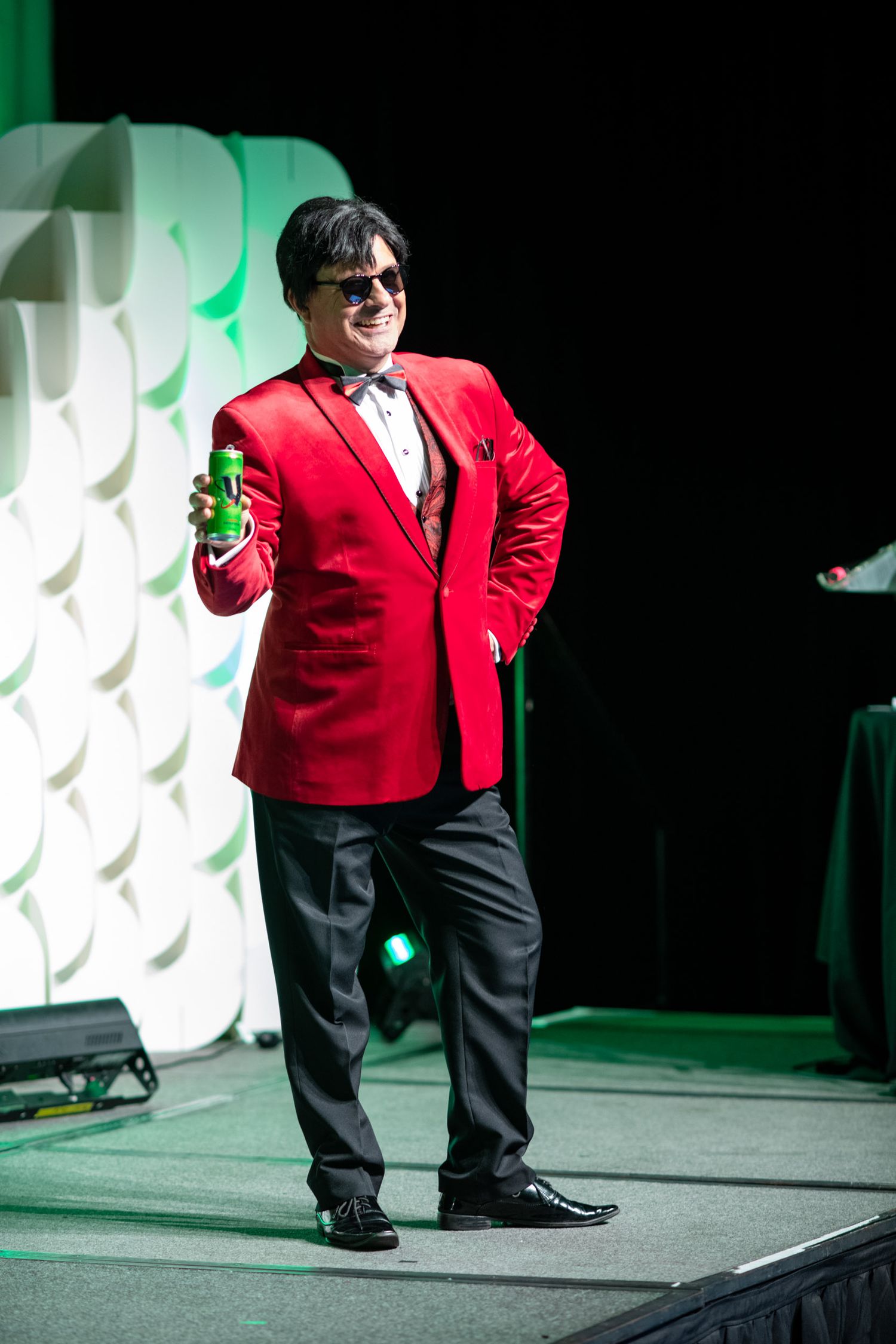 Man in red blazer and sunglasses holding green bottle on stage.