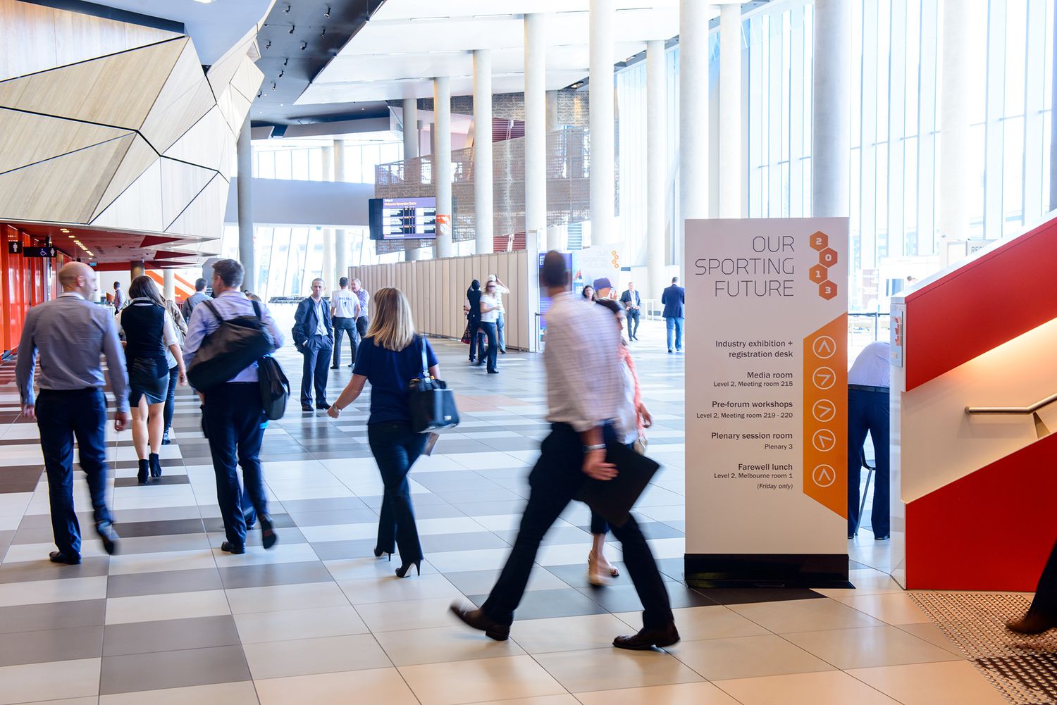 Business professionals walking through a modern convention center near an Our Sporting Future banner.