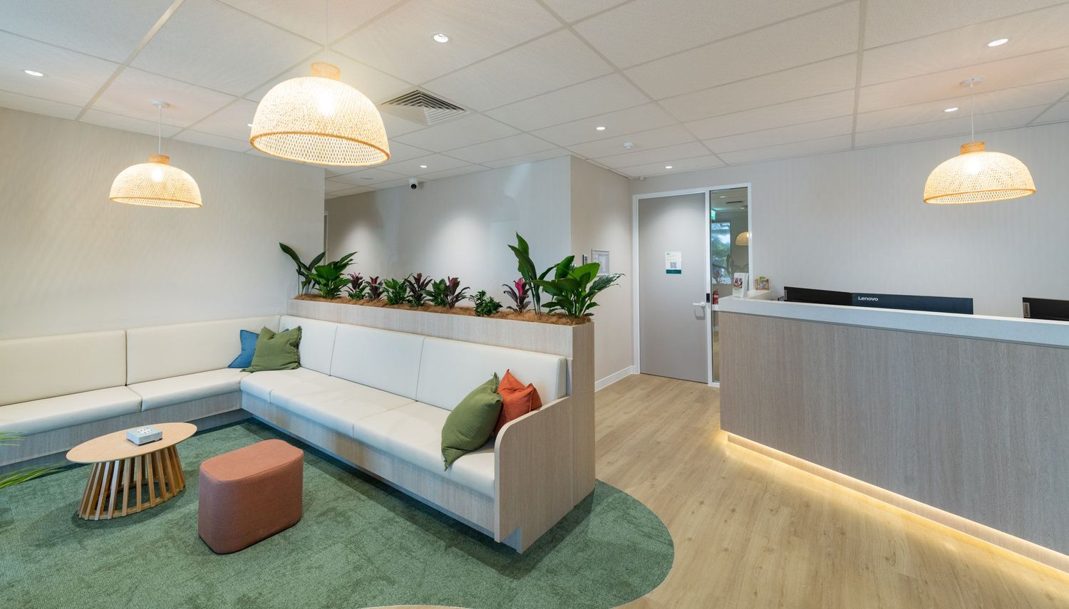 Bright modern office lounge with white sofa, teal rug, pendant lights, reception desk, and decorative indoor plants.
