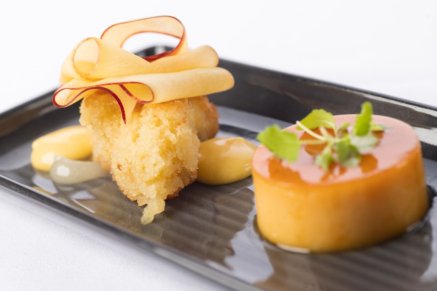 Upscale dessert duo featuring crispy fried cake with ribbon garnish and glazed tart on black plate.