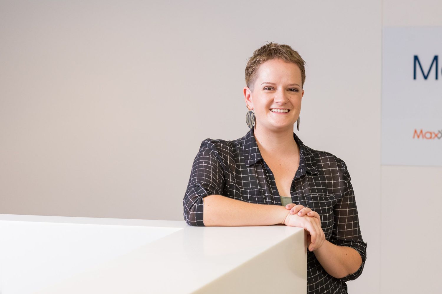 Smiling woman with short hair in patterned blazer leans on white reception desk in a bright modern office.