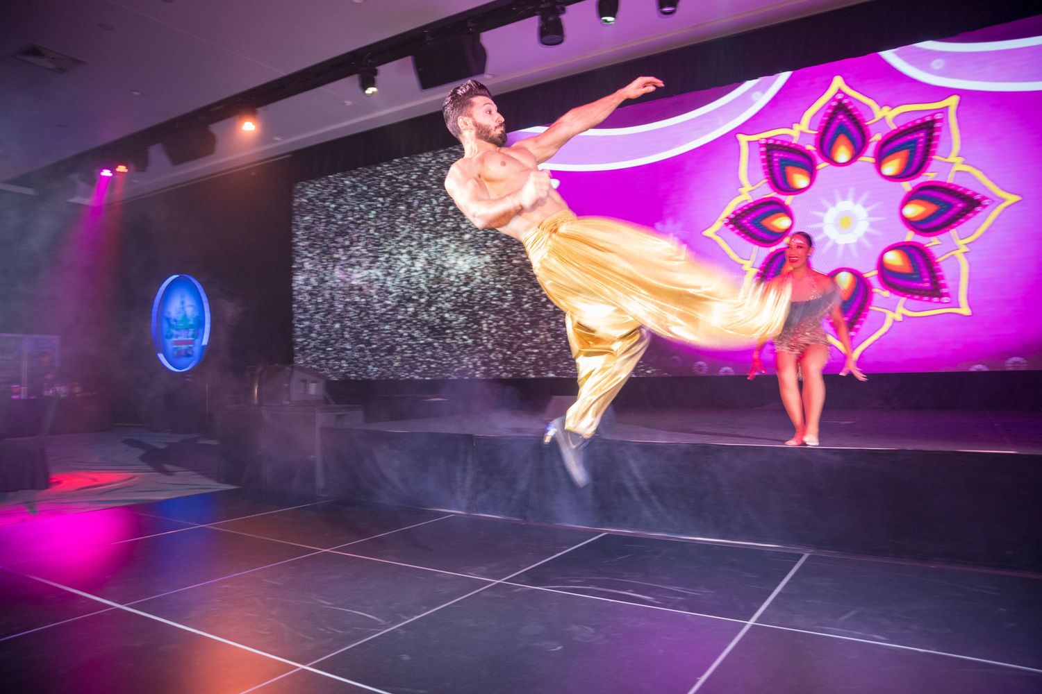 Male dancer leaping gracefully in gold costume on stage with a floral mandala backdrop.