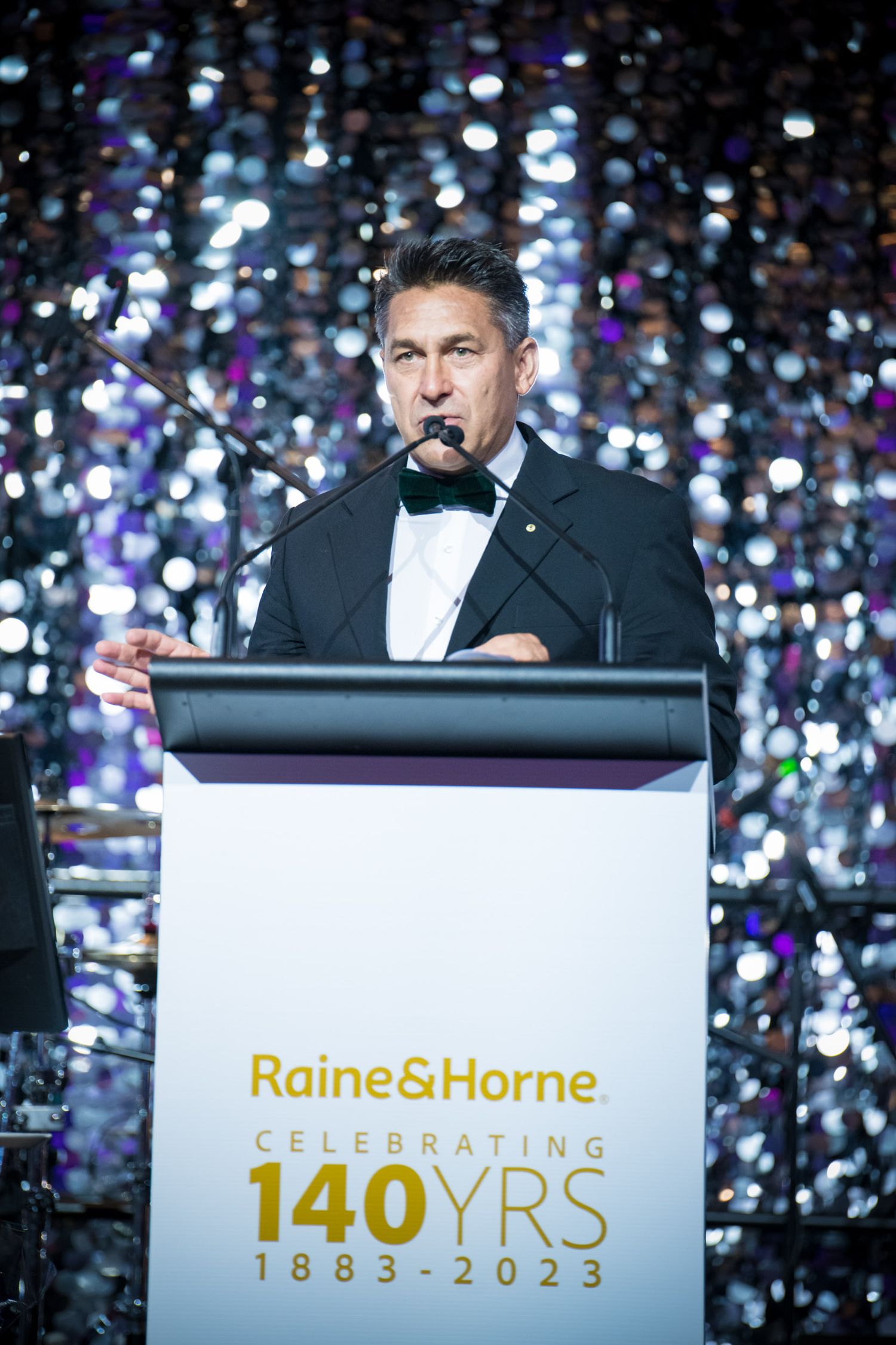 Man in tuxedo speaks at podium celebrating Raine and Horne 140 years anniversary gala event 1883 to 2023.