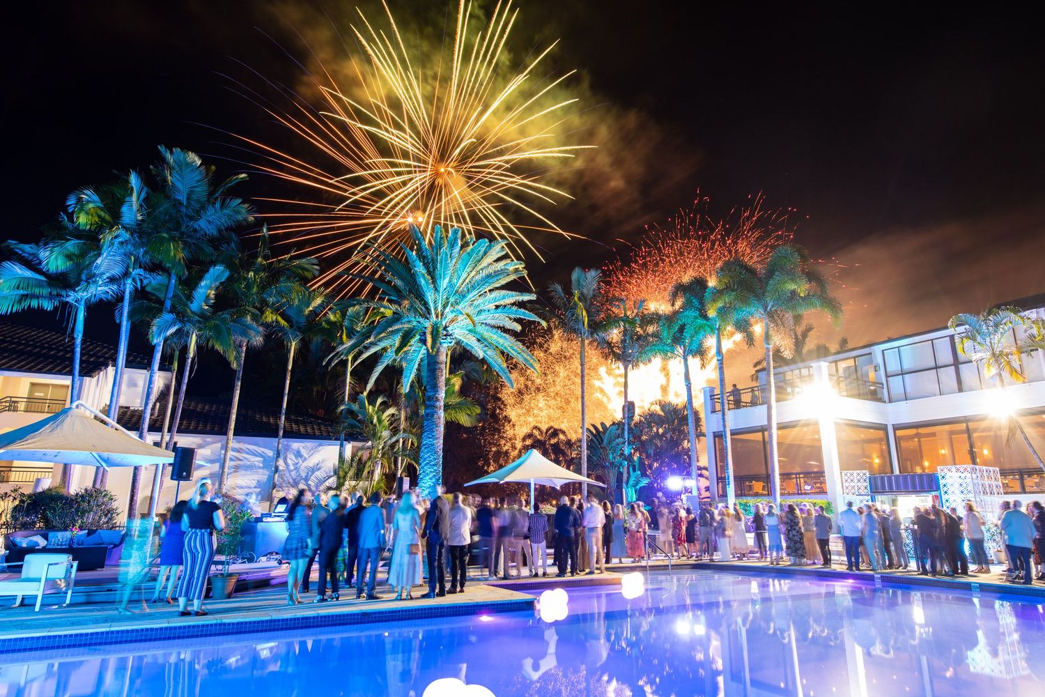 Outdoor pool party at night with colorful fireworks exploding over palm trees and guests gathered around.