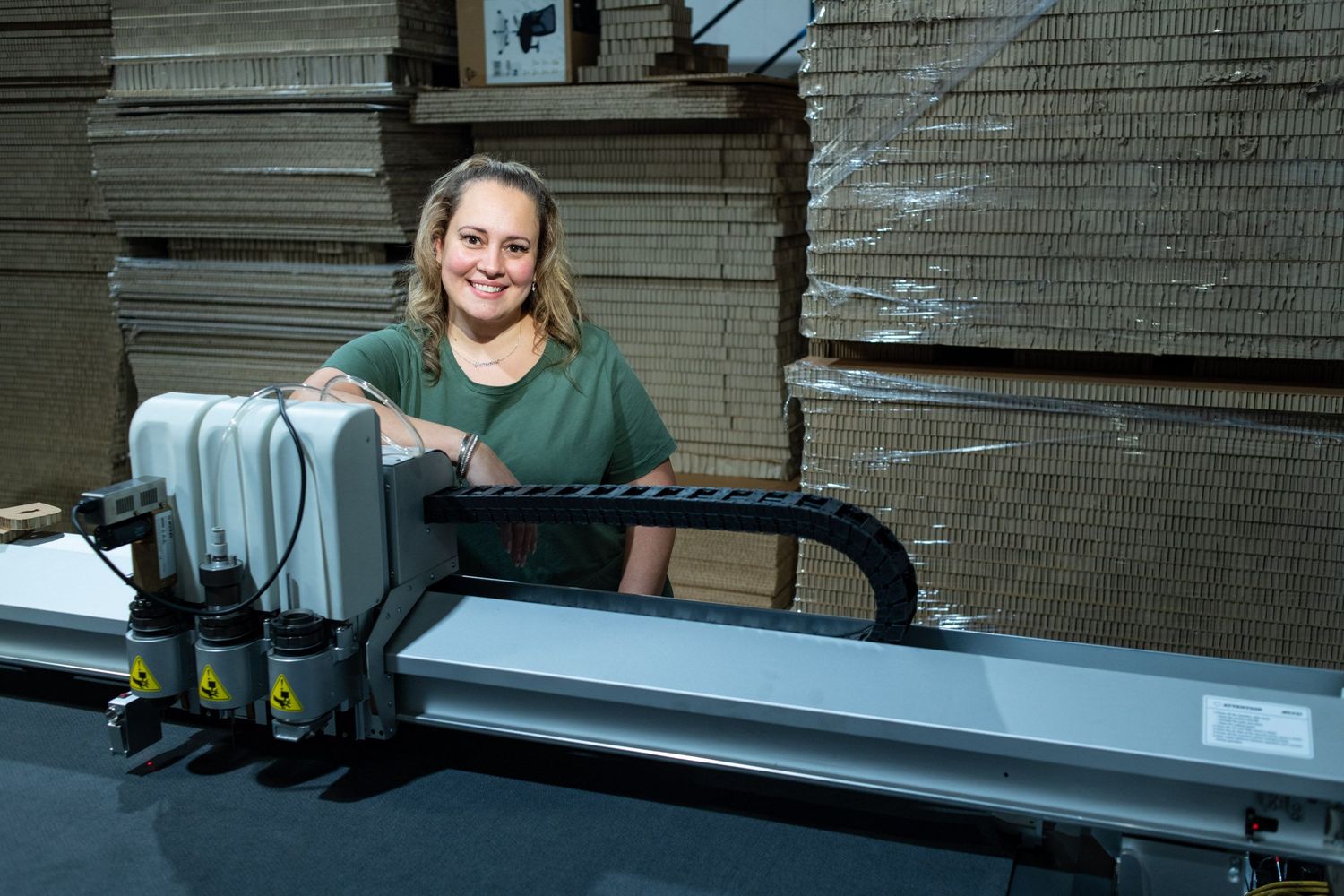 Smiling woman operating a large cutting machine surrounded by stacked cardboard in a warehouse.