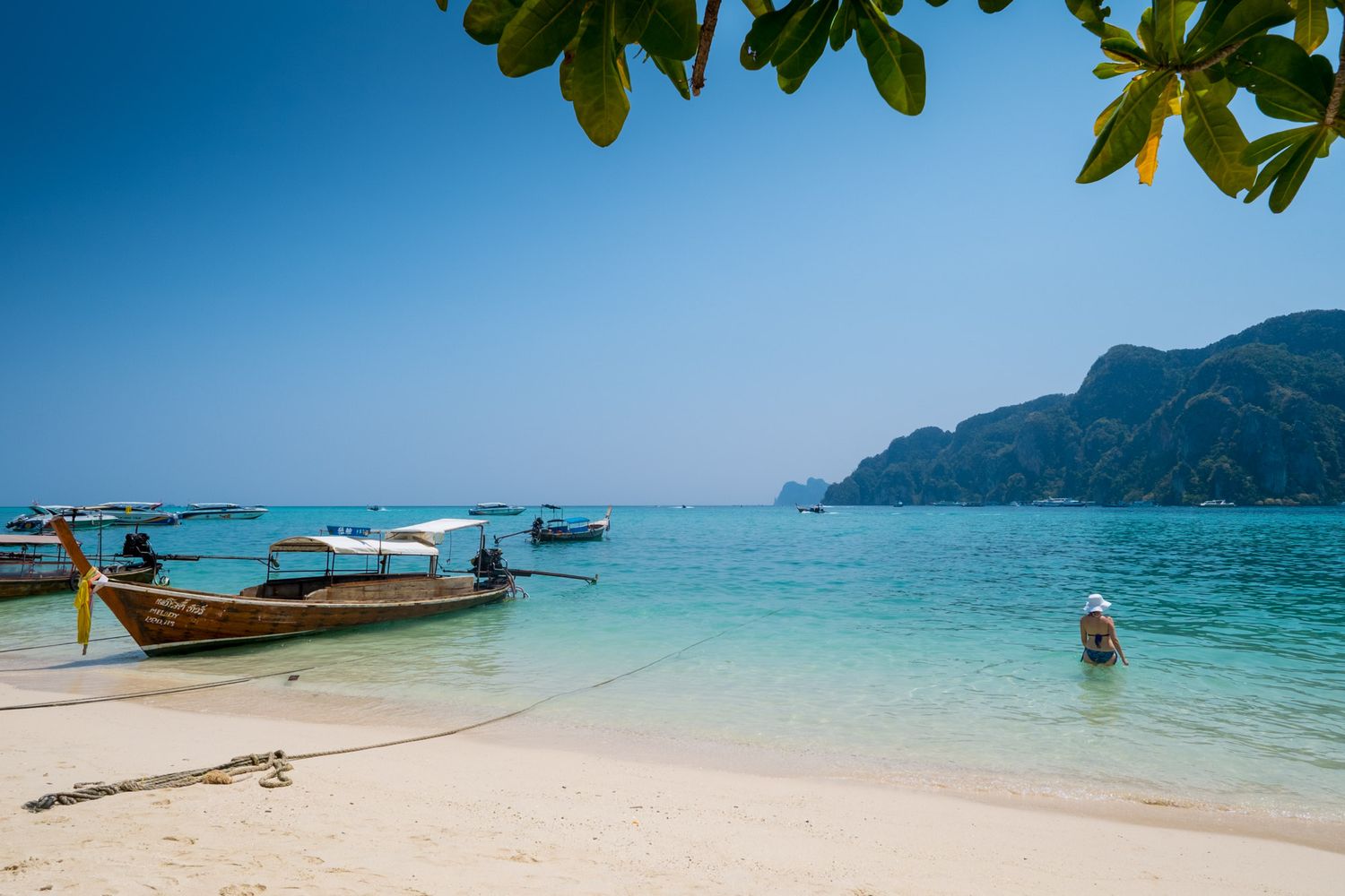 Longtail boats float near a white sand beach with clear turquoise water and limestone cliffs in Thailand.
