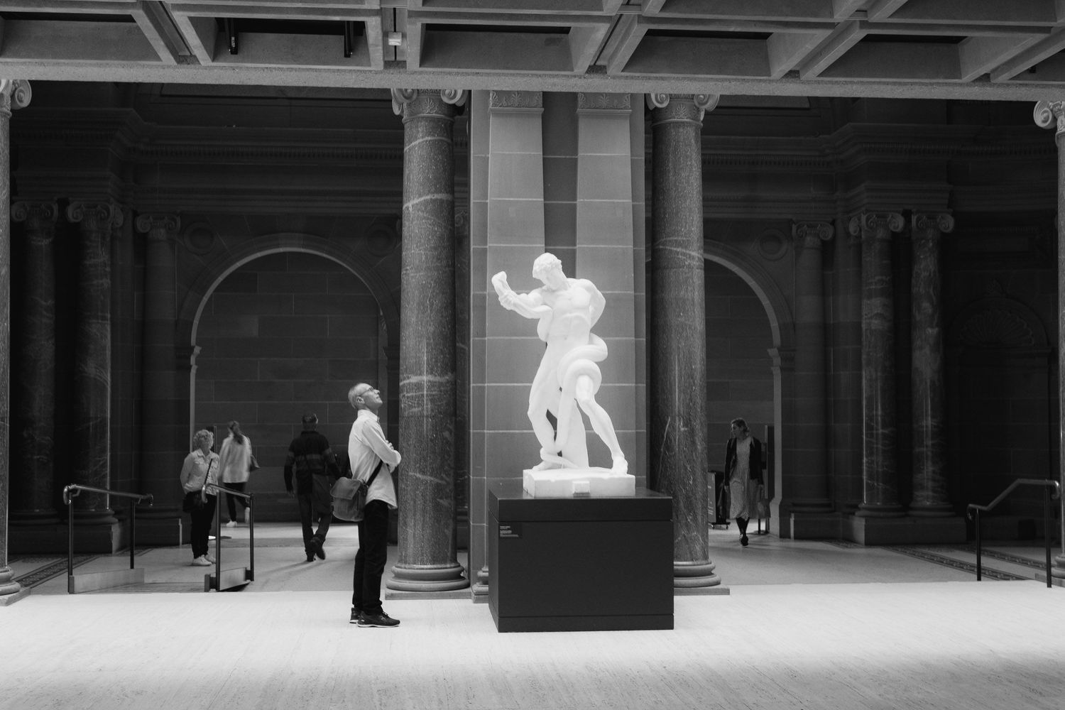 Black and white photo of visitors admiring a classical marble sculpture in a grand museum gallery.