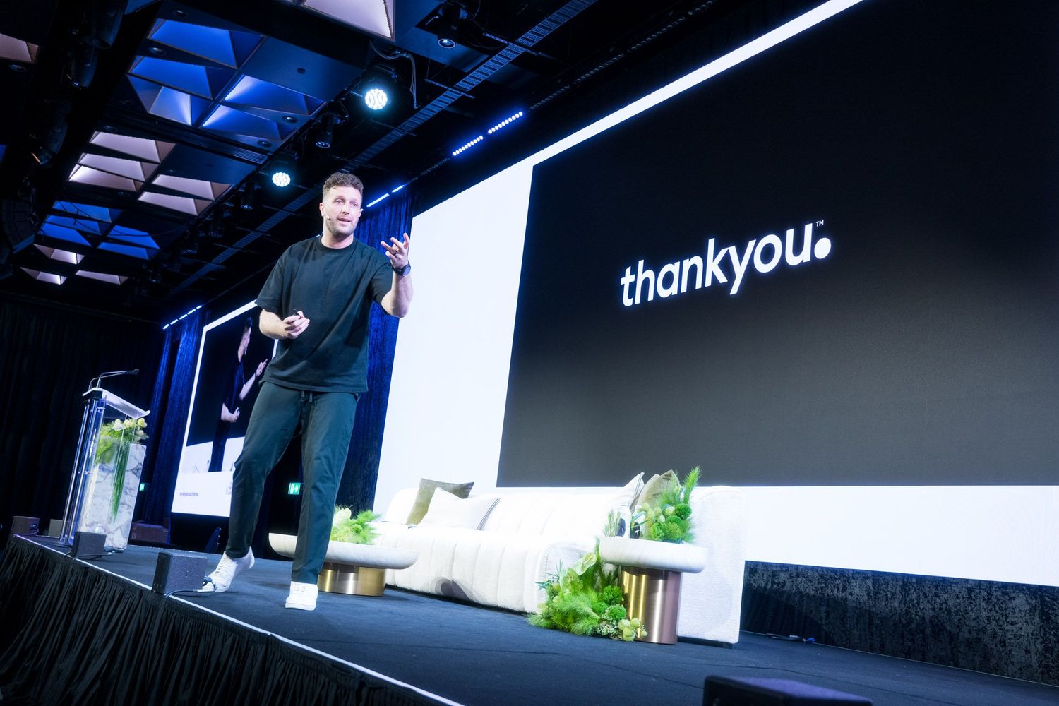 Male presenter on dark stage with thankyou brand logo displayed on large screen behind.
