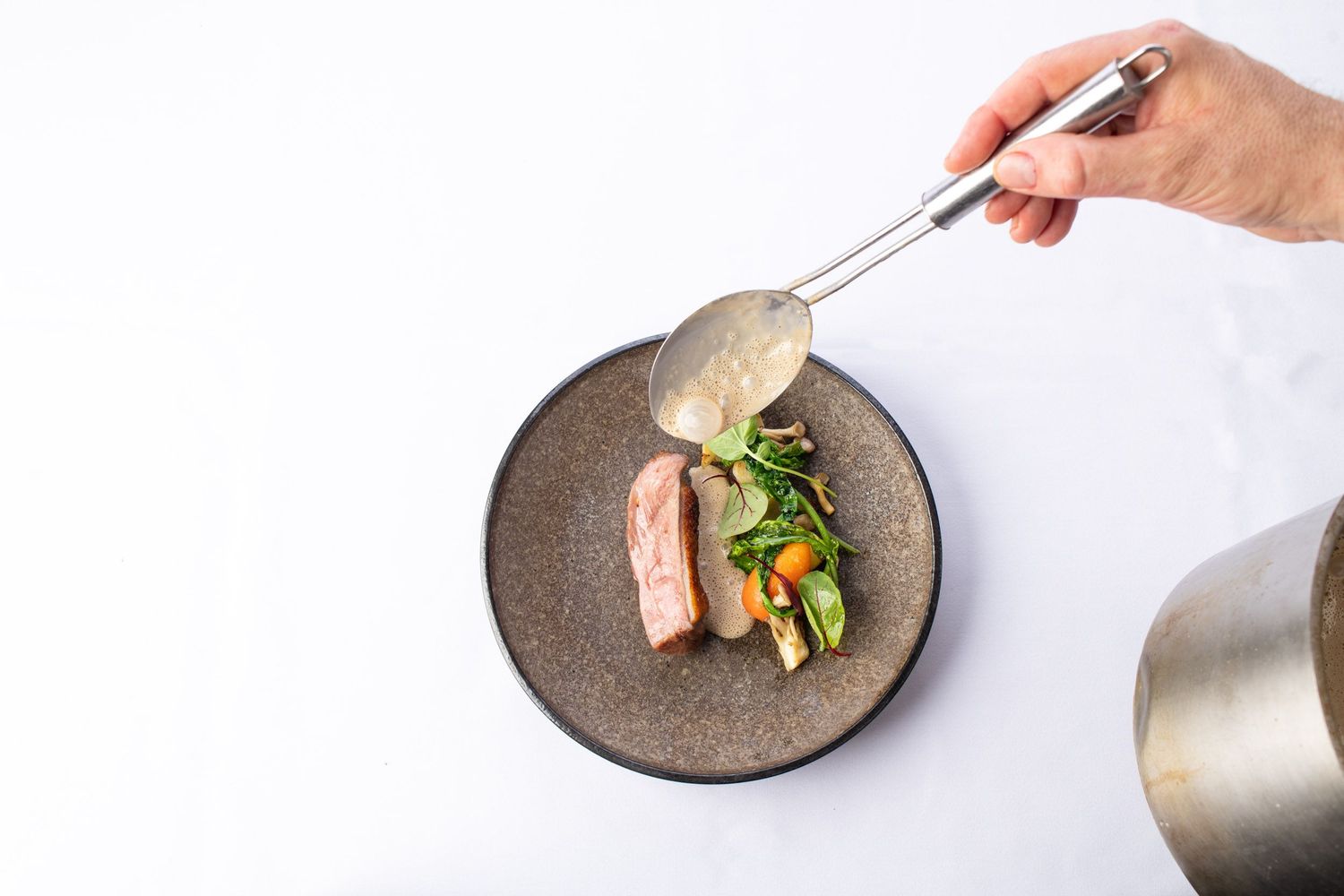 Chef pouring sauce over a gourmet meat dish with vegetables on a rustic ceramic plate.