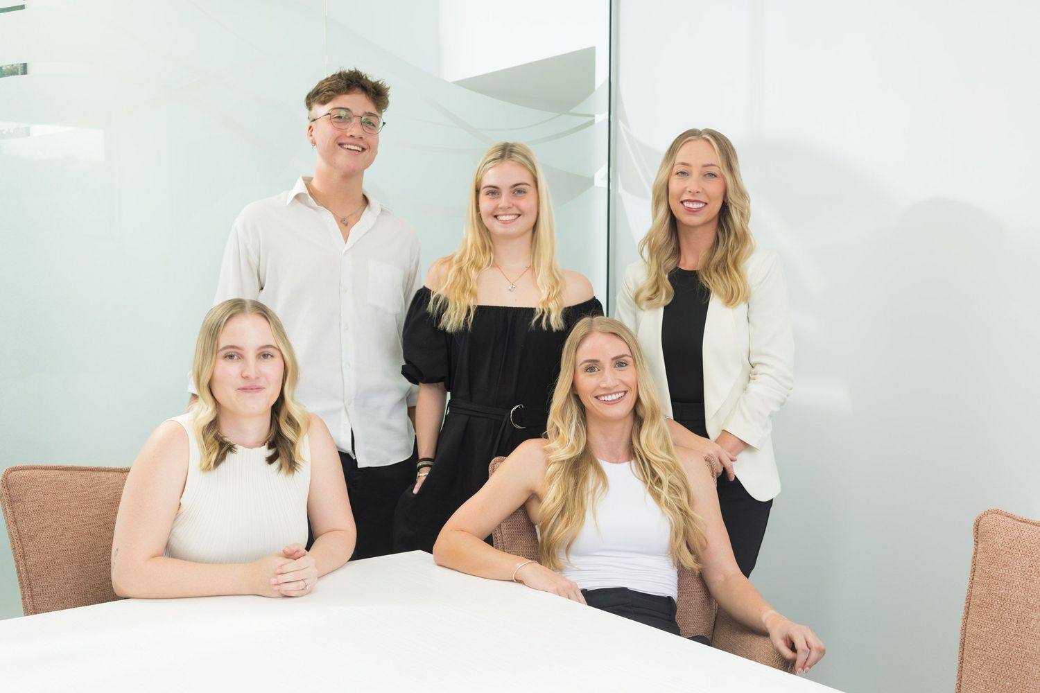 Five young professionals smiling together in a bright modern office conference room.