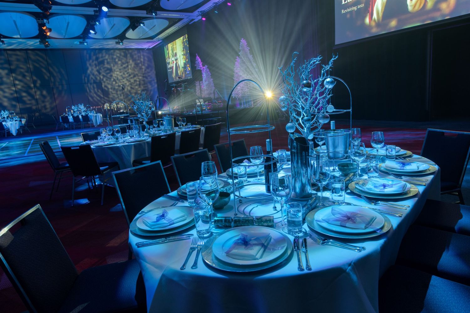 Elegantly set oval dining table with white linens, fine glassware, and decorative centerpiece at a blue-lit gala.