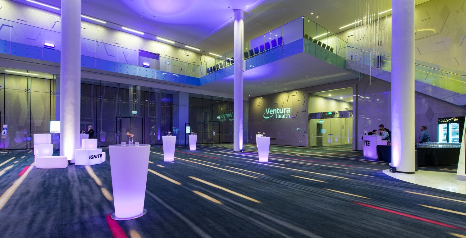 Spacious modern event venue lobby with purple ambient lighting, glowing white vases and carpeted floors.