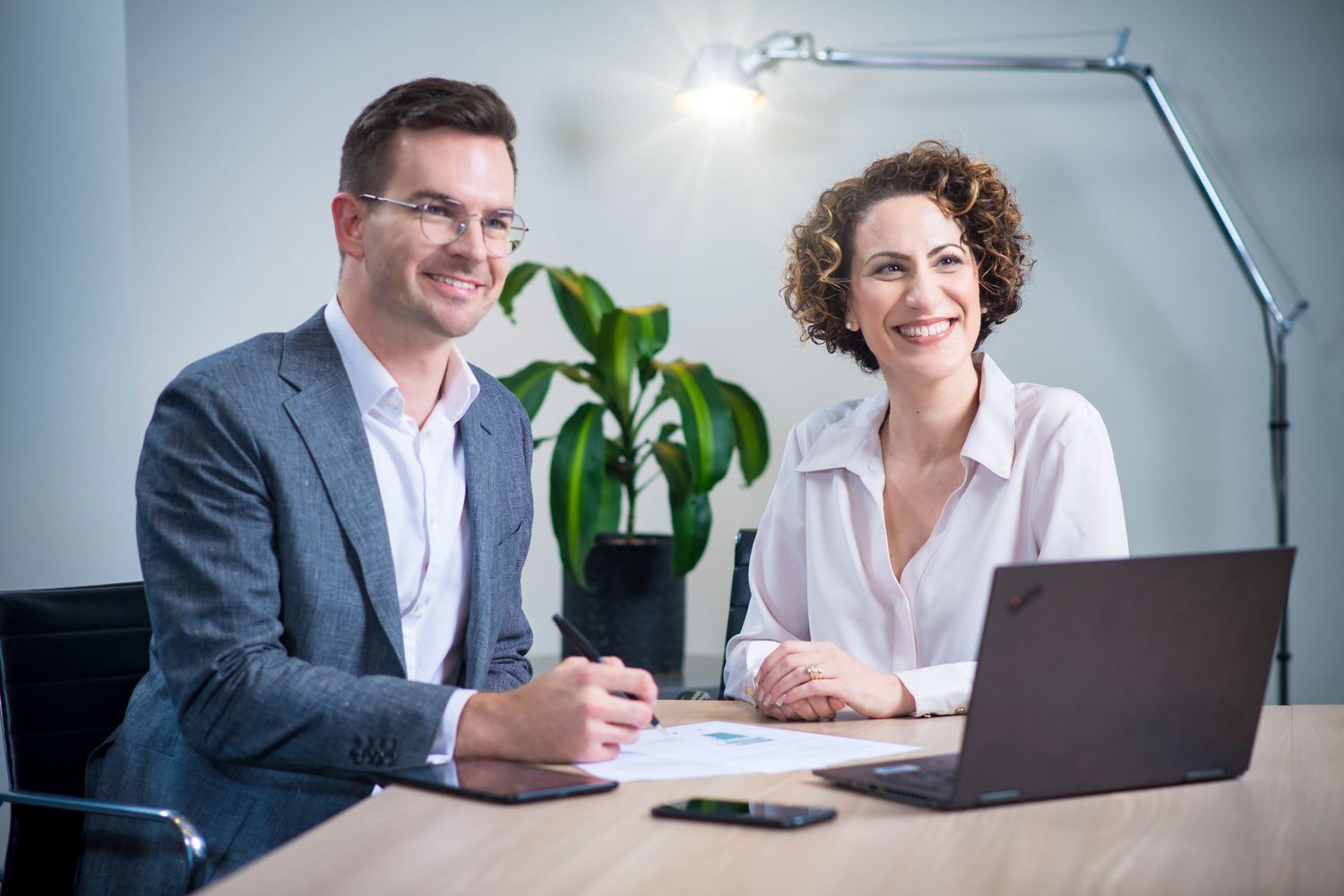 Man and woman smiling at office desk with laptop in bright workspace.