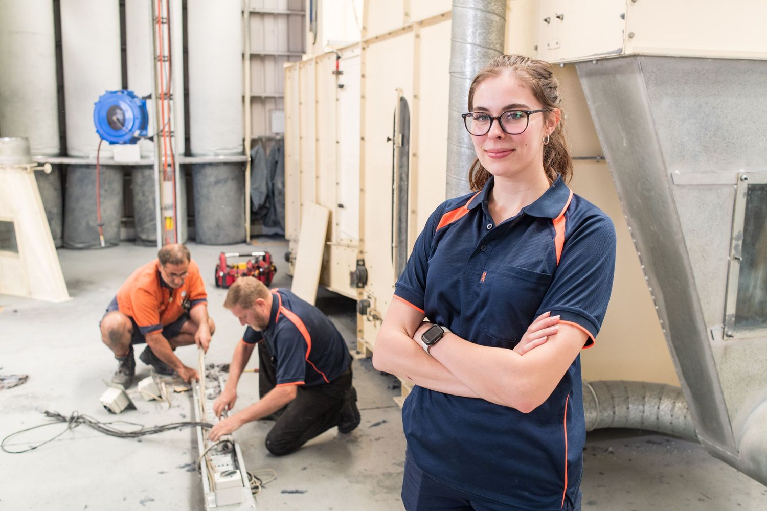 Young woman in blue workwear standing with crossed arms in an industrial workspace with colleagues.