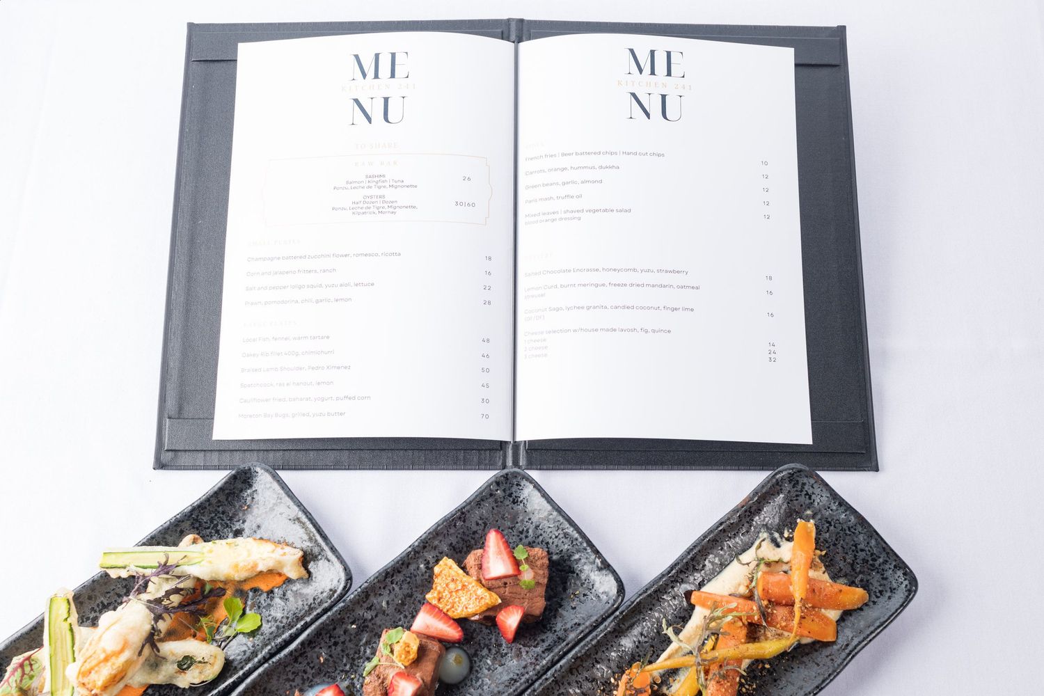 Open restaurant menu book displayed above three dark plates of elegant gourmet appetizers.