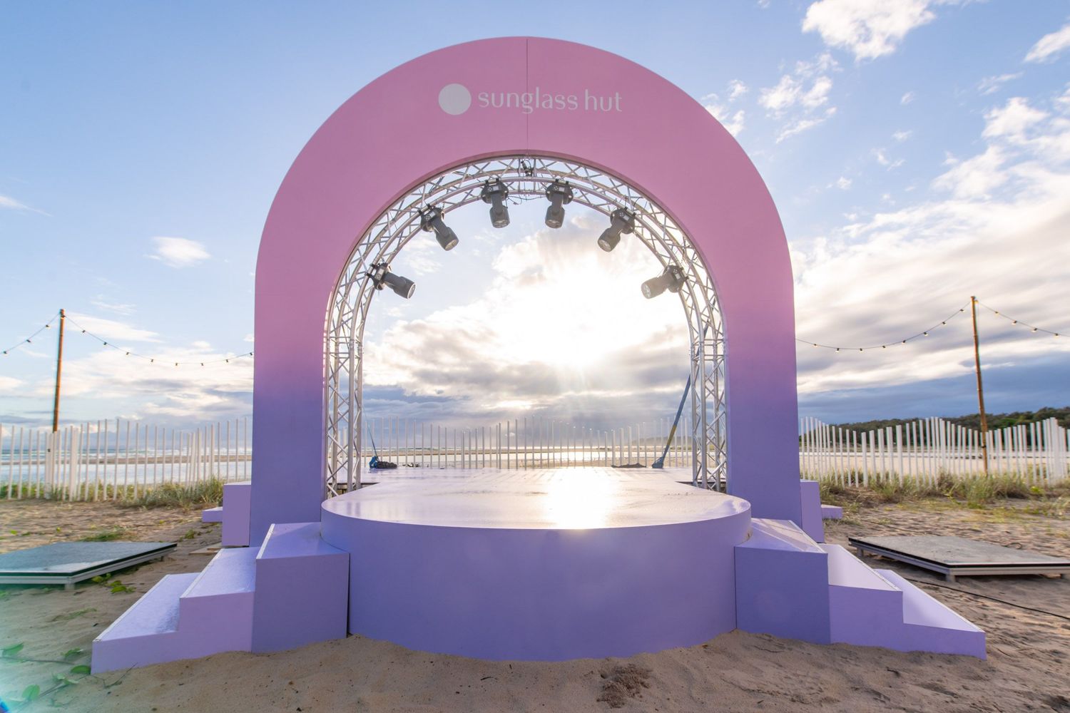 Pink and purple gradient arch stage setup on beach at sunset with spotlights.