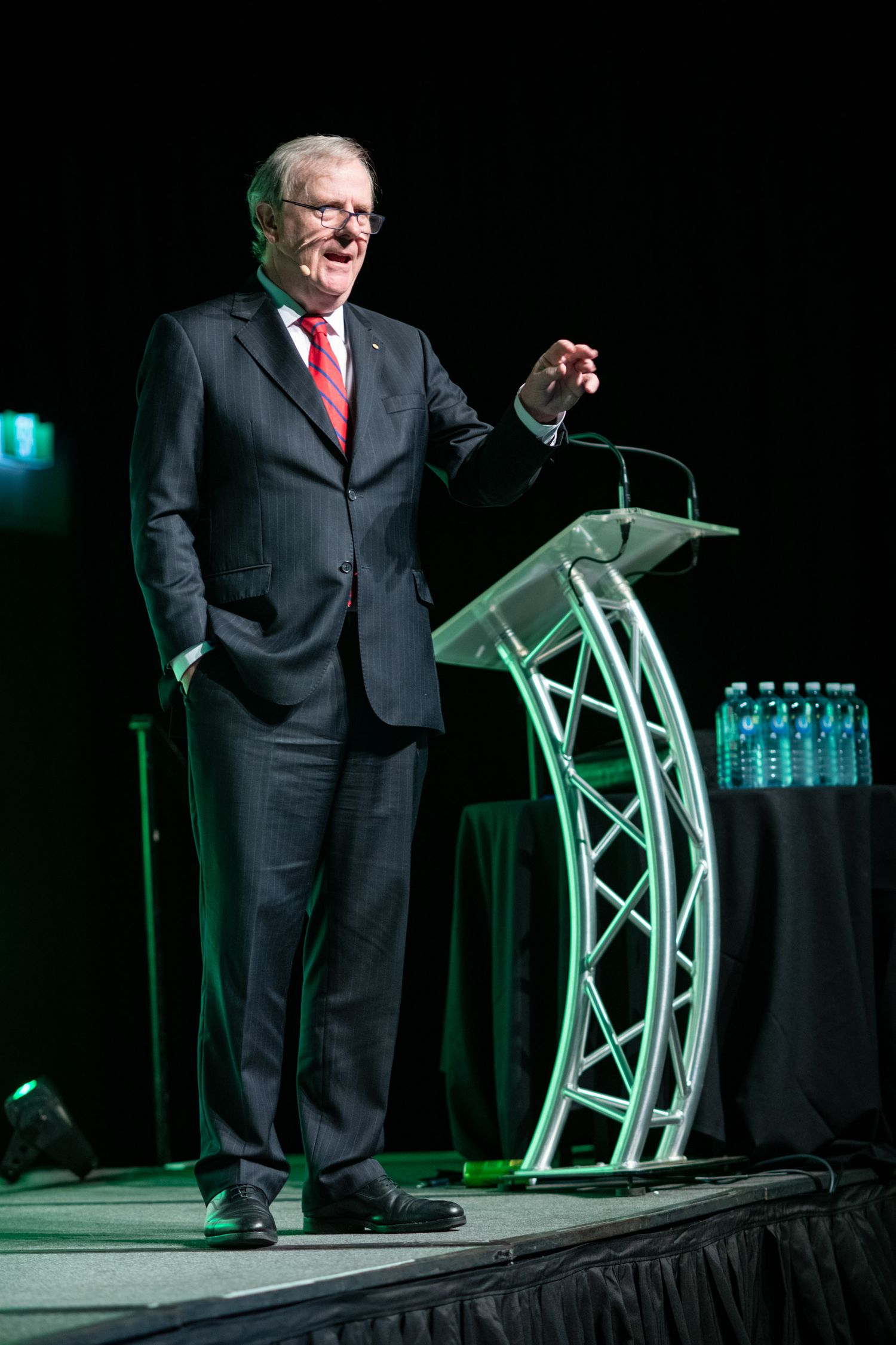 Senior businessman in dark suit delivering speech at modern glass podium on a dimly lit stage.
