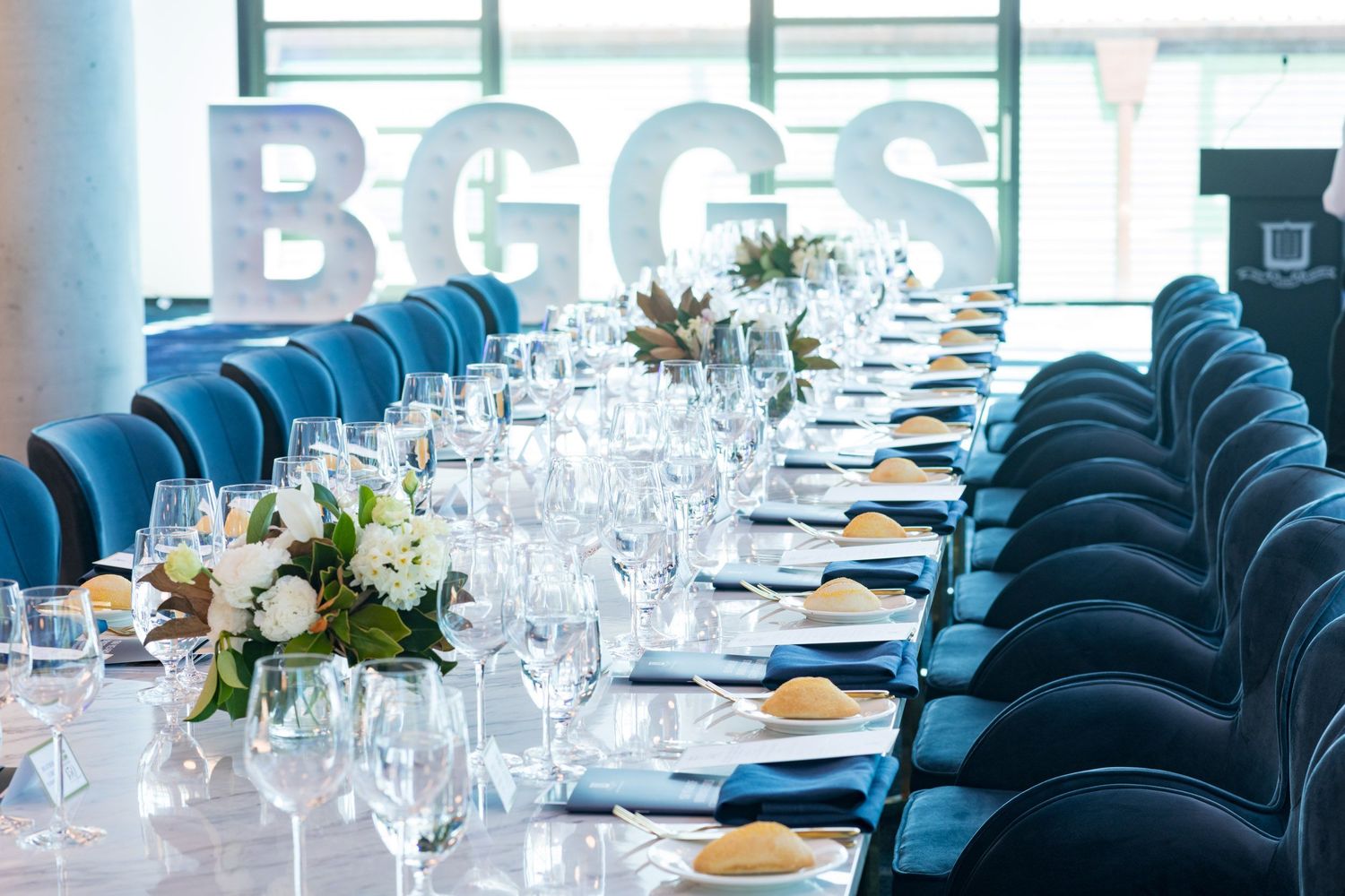 Elegant long dining table set for a formal gala event with blue velvet chairs and large BGGS letters in the background.