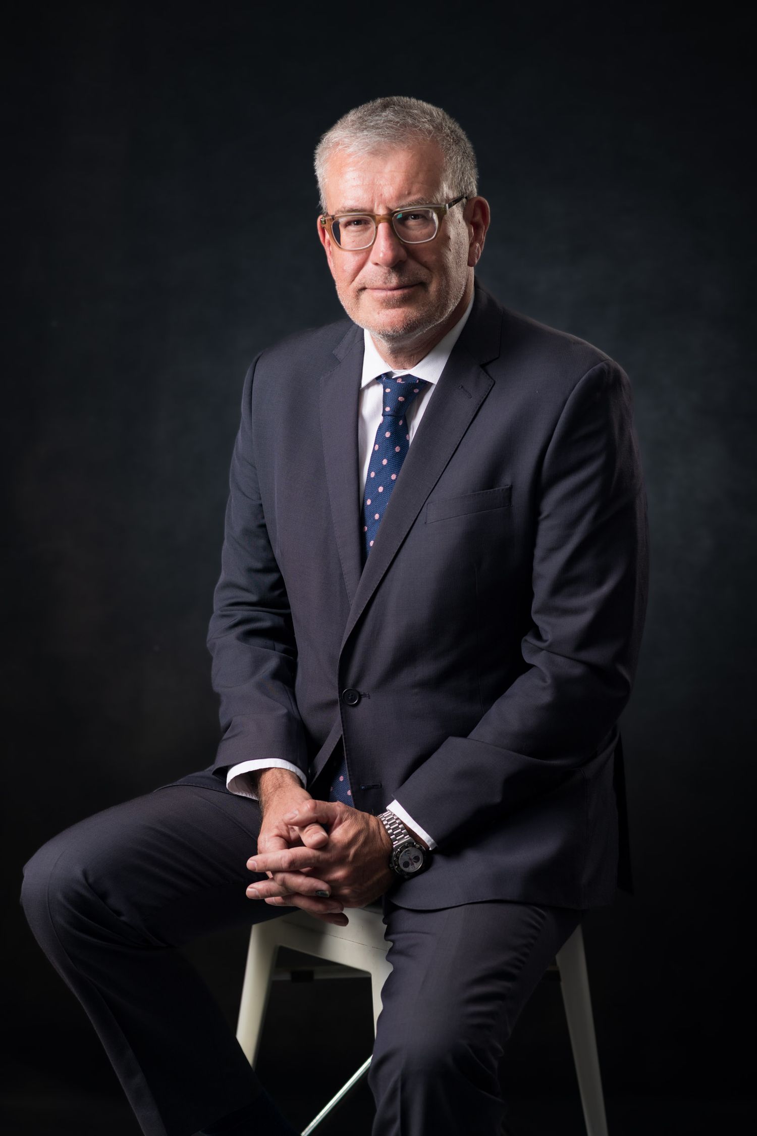 Professional businessman in dark suit seated on stool posing for corporate portrait on dark background.