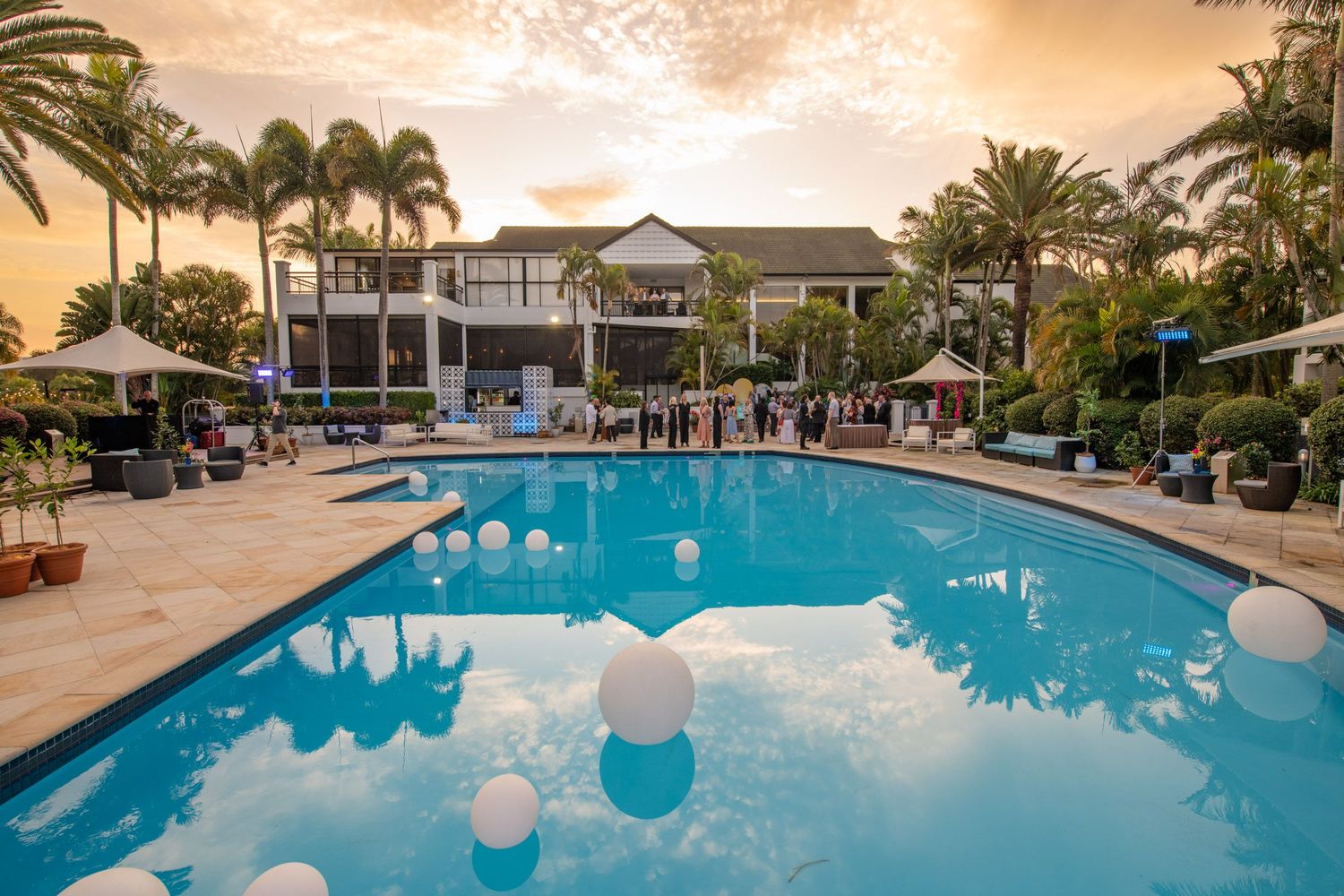Tropical resort pool at sunset decorated with floating white balloons and guests mingling at outdoor event.