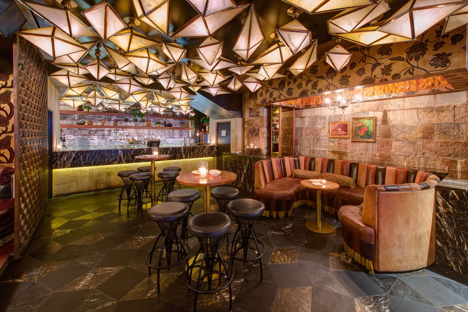 Stylish bar interior with geometric ceiling, leather booth seating, round tables, and warm ambient lighting.