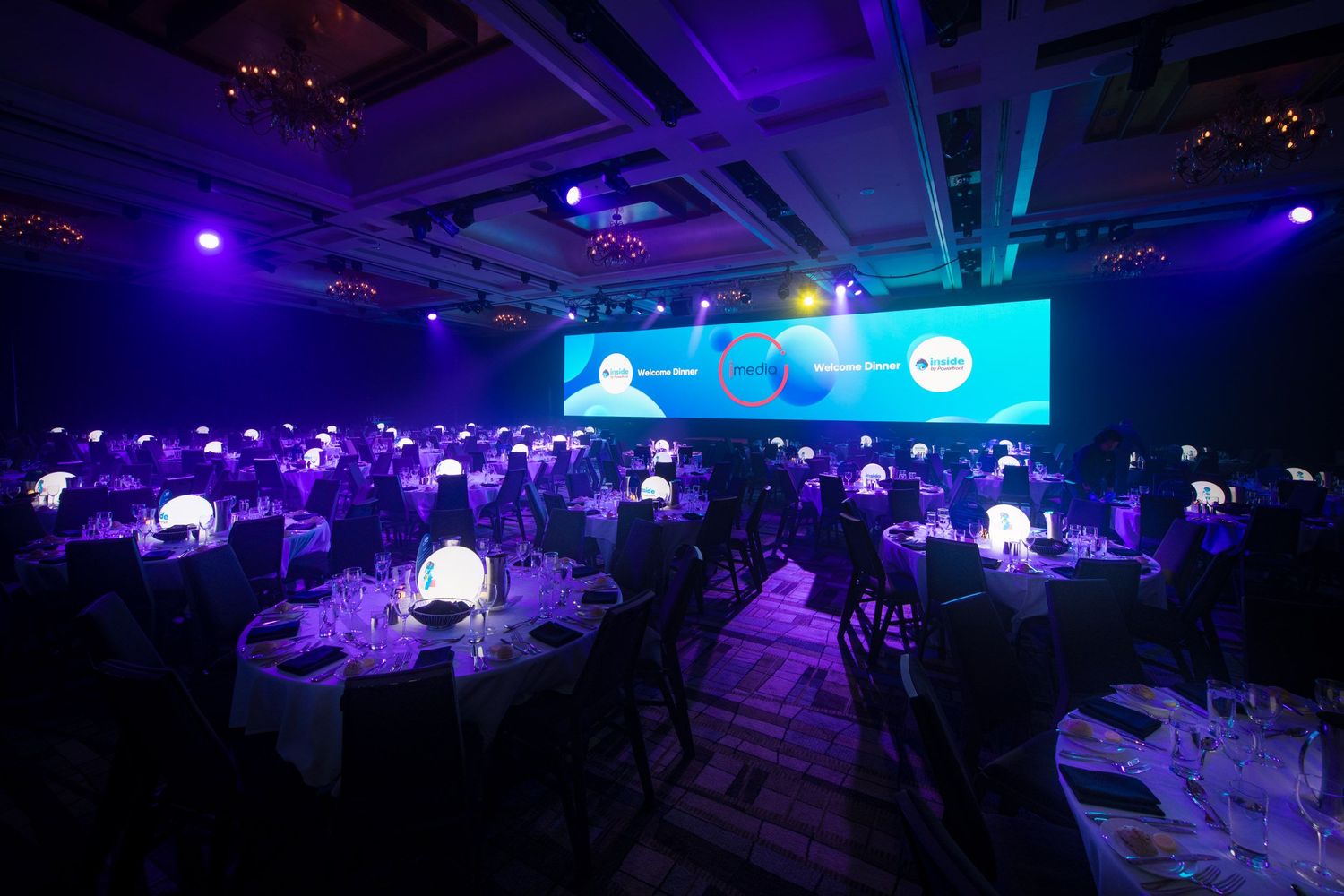 Elegant corporate gala dinner with round tables, blue lighting, and a large presentation screen in a ballroom.