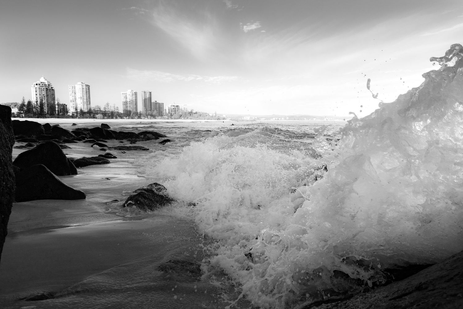 Black and white photo of powerful ocean wave crashing on rocks with city skyline.