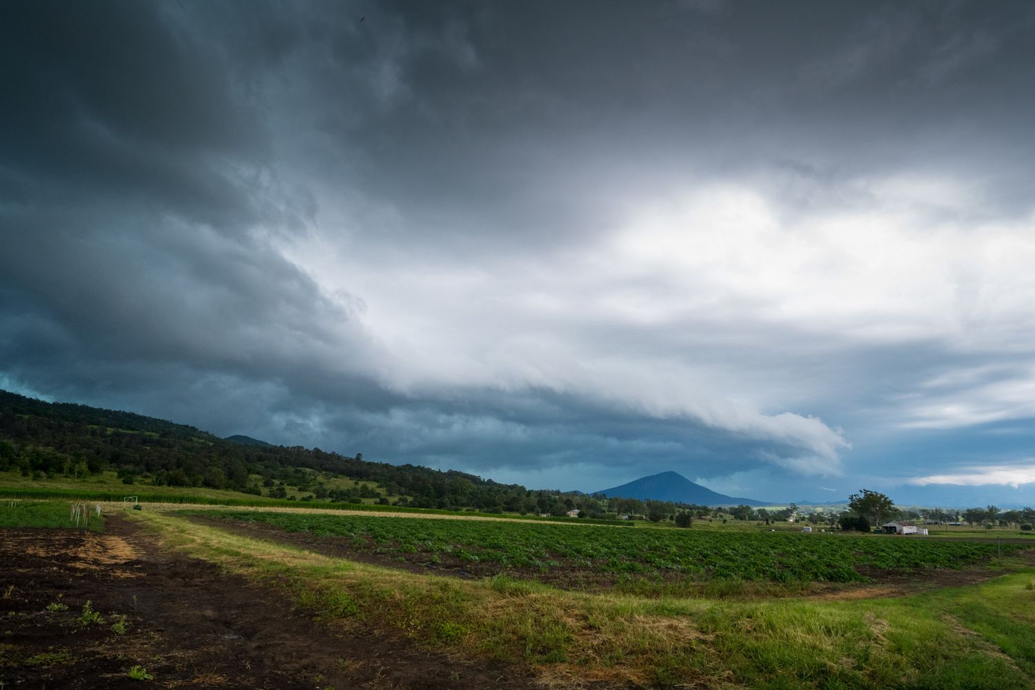 Storm clouds gather over lush green farmland with mountain backdrop in rural landscape.