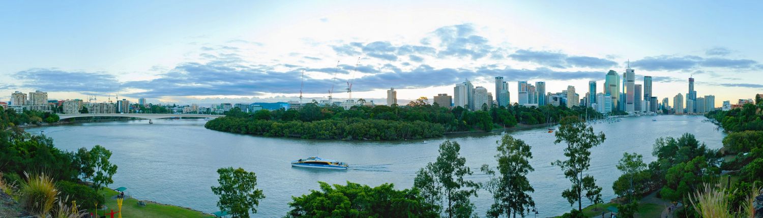 Panoramic view of Brisbane city skyline at dusk with the Brisbane River and lush green parklands in the foreground.