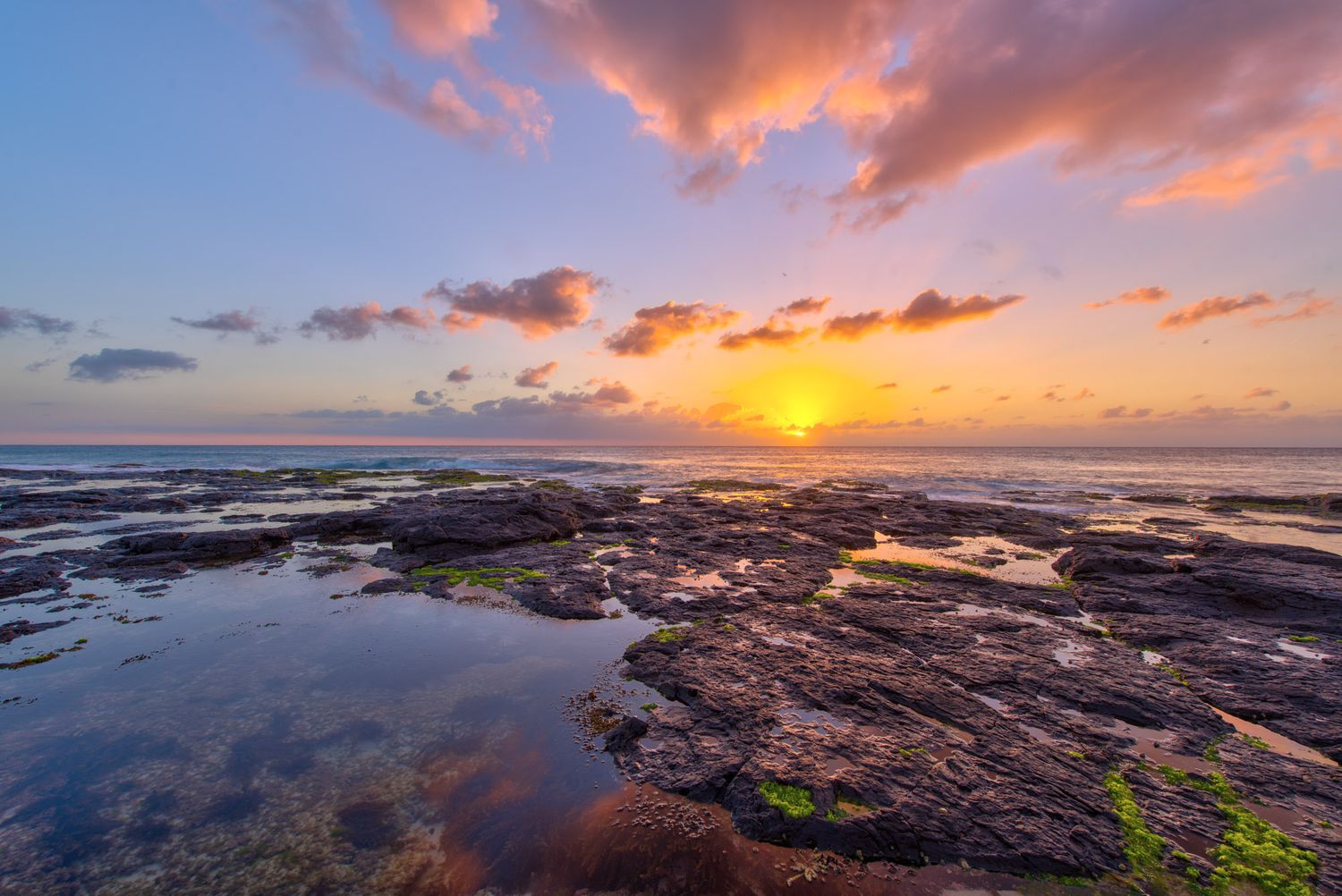 Stunning tropical sunset over rocky coastline with vibrant orange and pink clouds reflecting in tide pools.