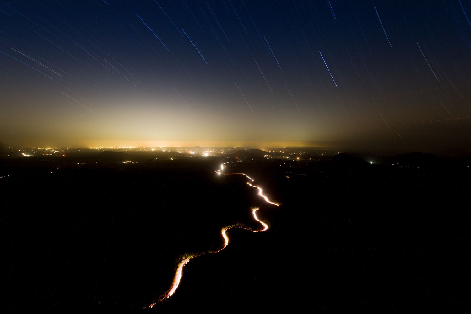 Long exposure night landscape showing star trails and glowing city lights with winding road below.